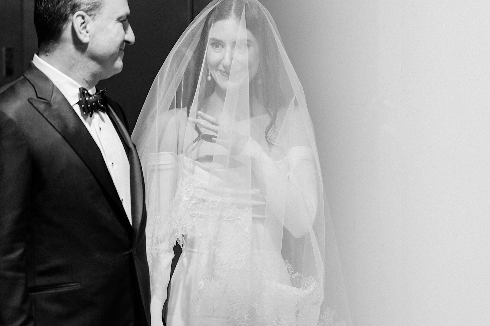 Bride in wedding dress and veil smiles at companion in tuxedo, hand to her chest. Black and white photograph with soft lighting captures their joy at the Kimmel Center on New Year's Eve.