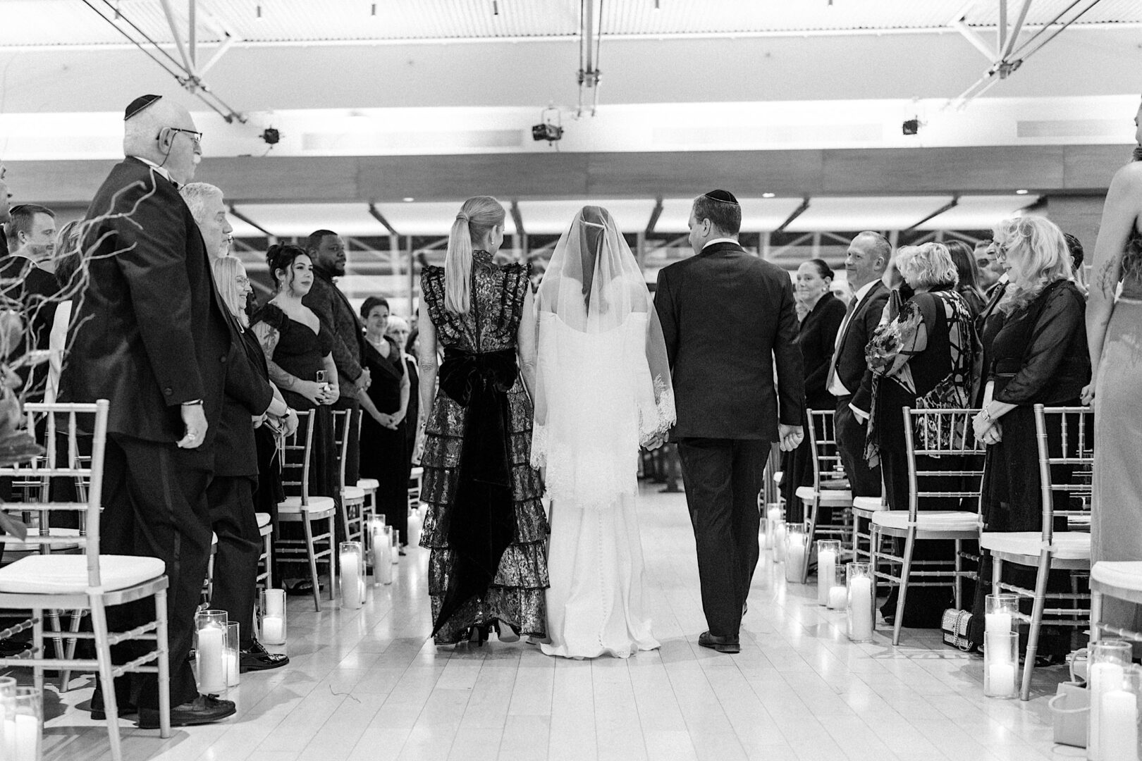A bride with a veil is walking down the aisle, escorted by two people, at the Kimmel Center in Philadelphia. Guests are seated on both sides, observing the beautiful New Year's Eve procession.
