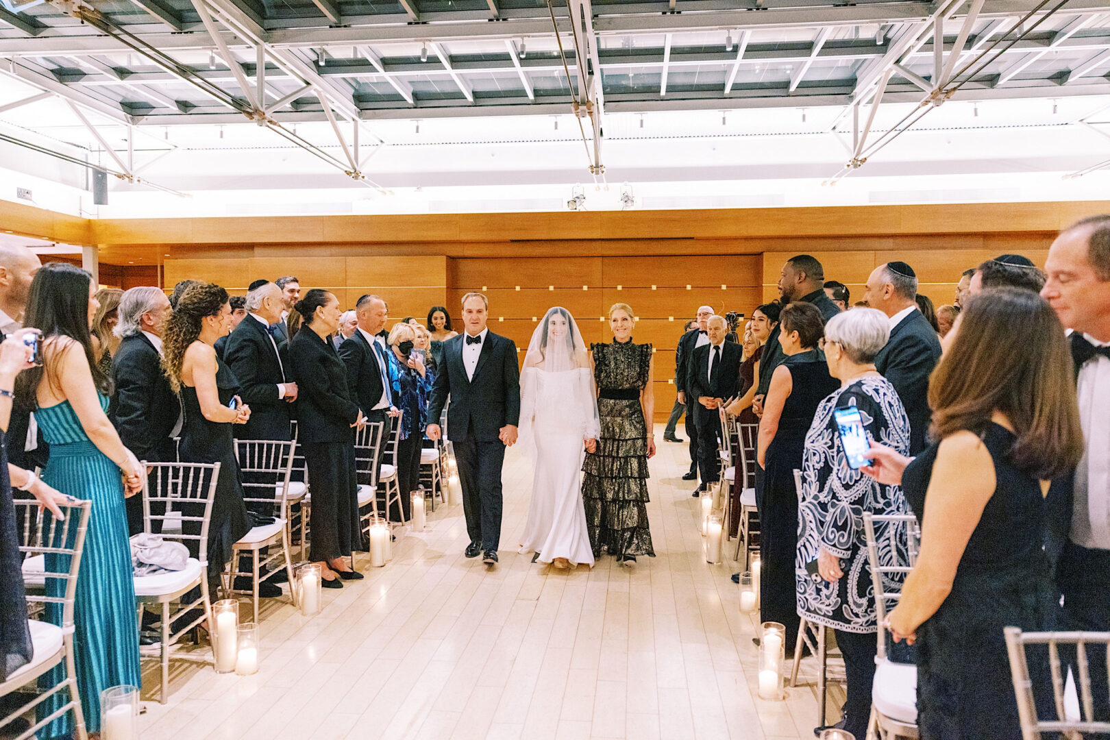 Bride walks down the aisle flanked by two people in a modern indoor venue at the Kimmel Center in Philadelphia, while guests stand on both sides taking photos and watching.