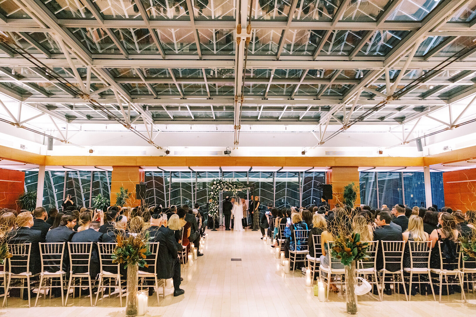 A wedding ceremony on New Year’s Eve in a modern, spacious hall in Philadelphia, with guests seated on white chairs, facing an altar adorned with flowers and greenery under a high glass ceiling.