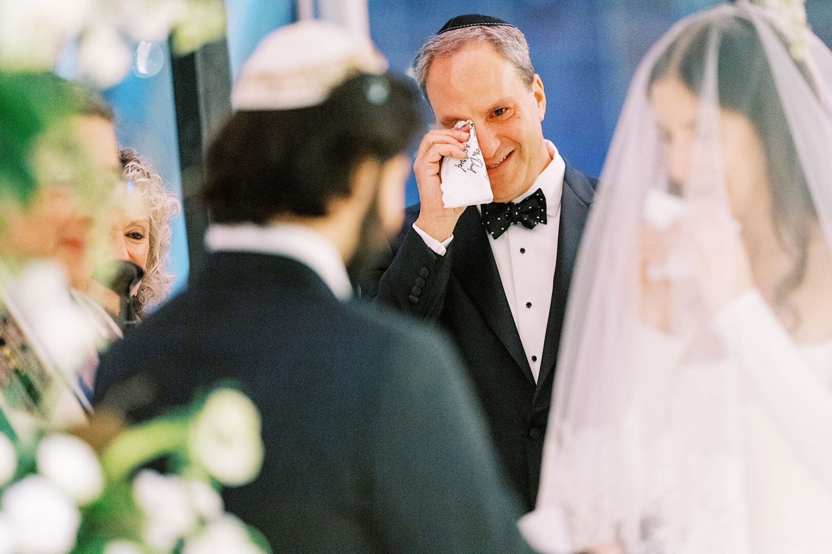 A man in a suit wipes tears with a handkerchief at a wedding ceremony. The bride in a veil and the groom are in the foreground, celebrating their special day at the Kimmel Center.