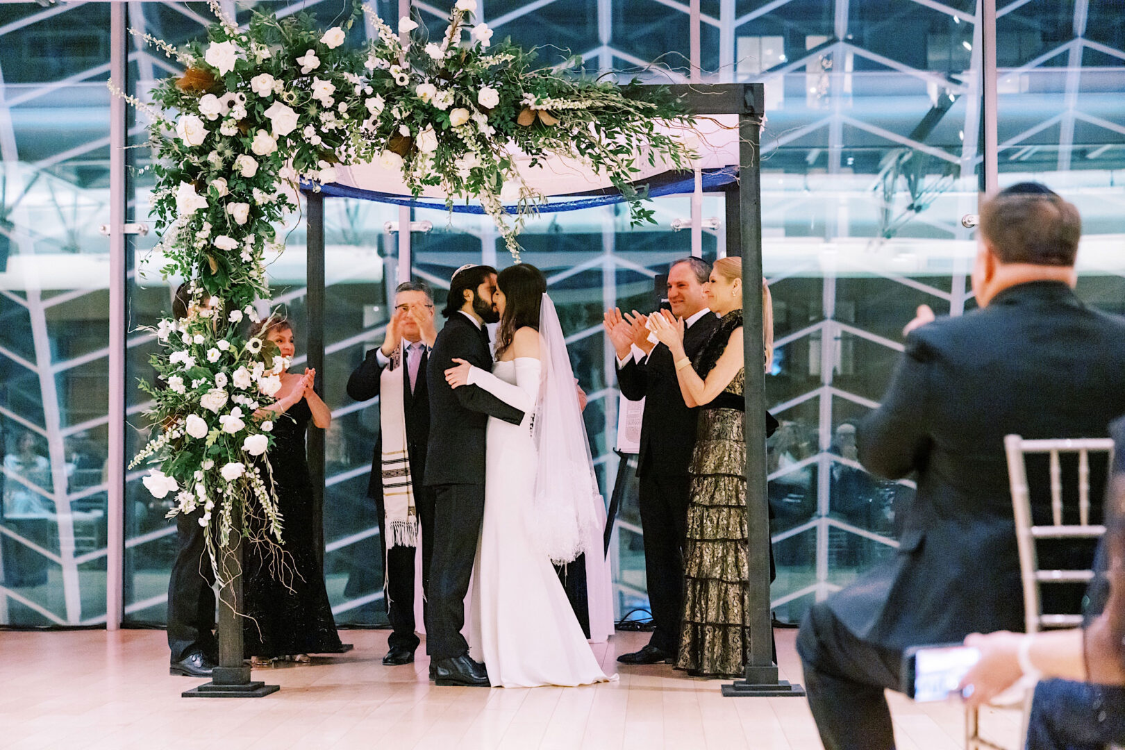 A couple, dressed in wedding attire, embraces under a floral-covered canopy with guests clapping and the elegant glass backdrop of the Kimmel Center in Philadelphia.