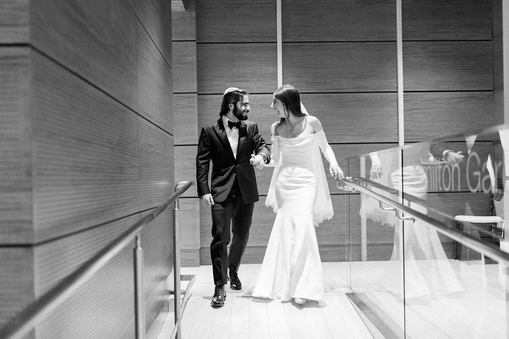 A couple dressed in formal attire, with the man in a suit and the woman in an off-the-shoulder gown, walk hand in hand down a modern hallway at the Kimmel Center, as if stepping into their own magical New Year's Eve celebration.