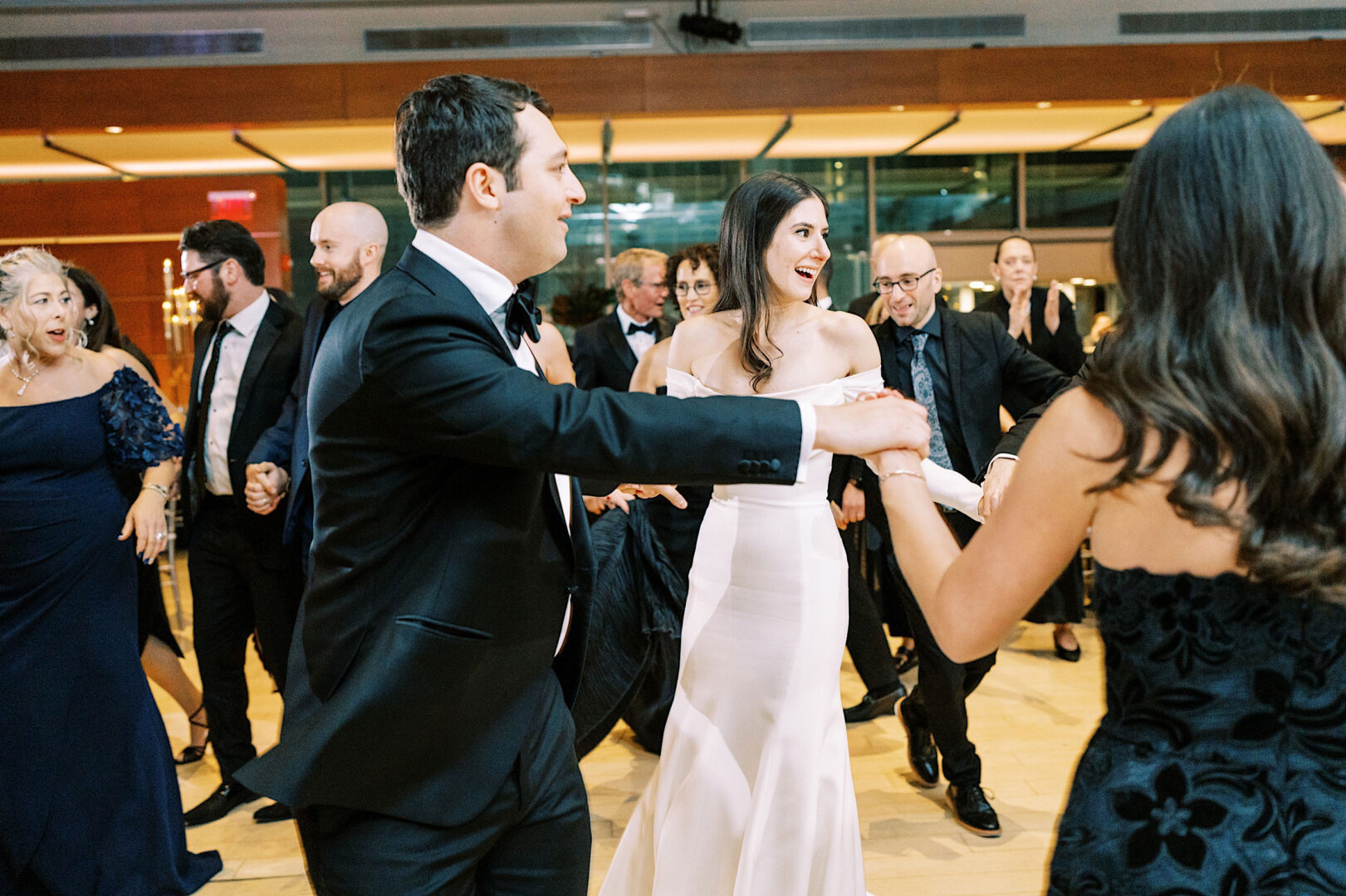 A bride and groom in formal attire dance with guests in a well-lit indoor venue, surrounded by other elegantly dressed attendees, celebrating their wedding in Philadelphia.