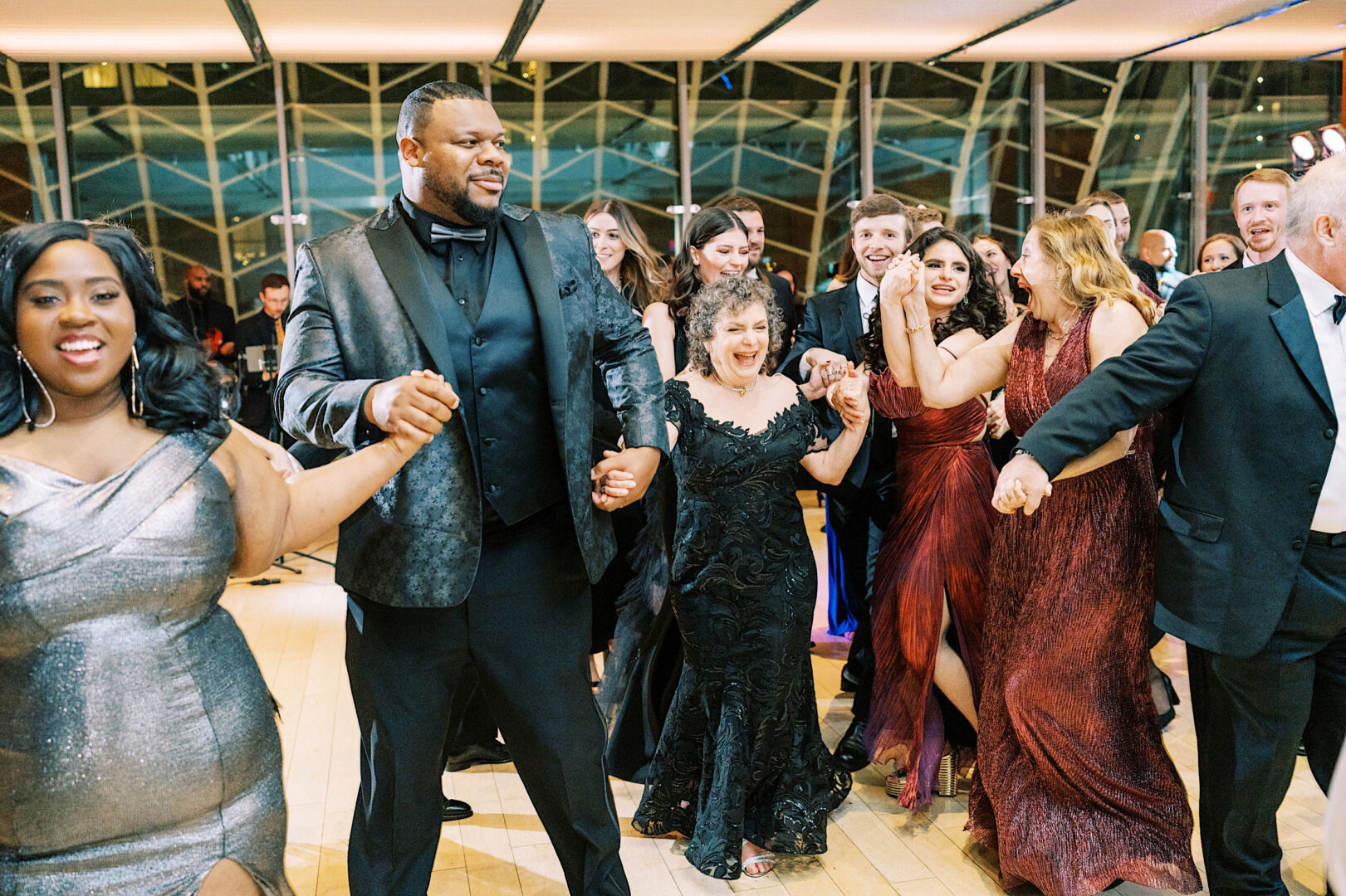 A group of people dressed in formal attire are holding hands and dancing joyfully in a well-lit room with large windows, celebrating a beautiful wedding at the Kimmel Center in Philadelphia.