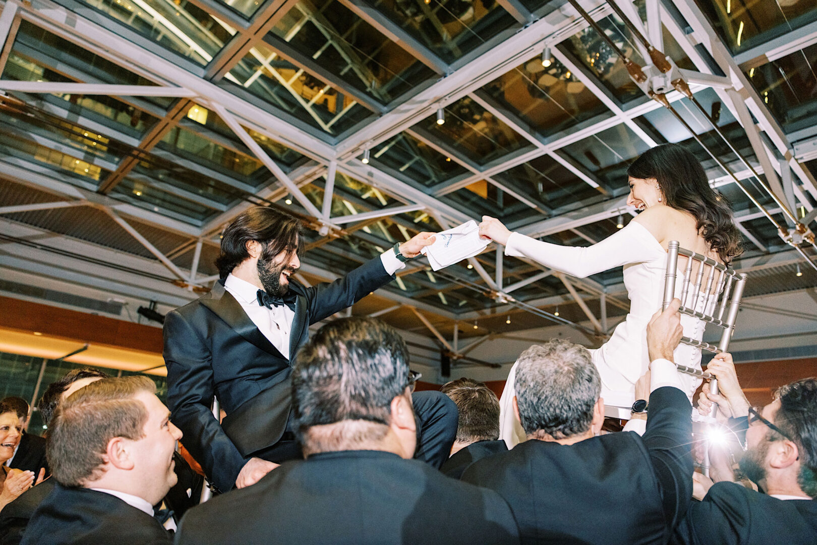 During a wedding celebration at the Kimmel Center in Philadelphia, a bride and groom are lifted on chairs by guests in a well-lit indoor venue with a stunning glass ceiling.
