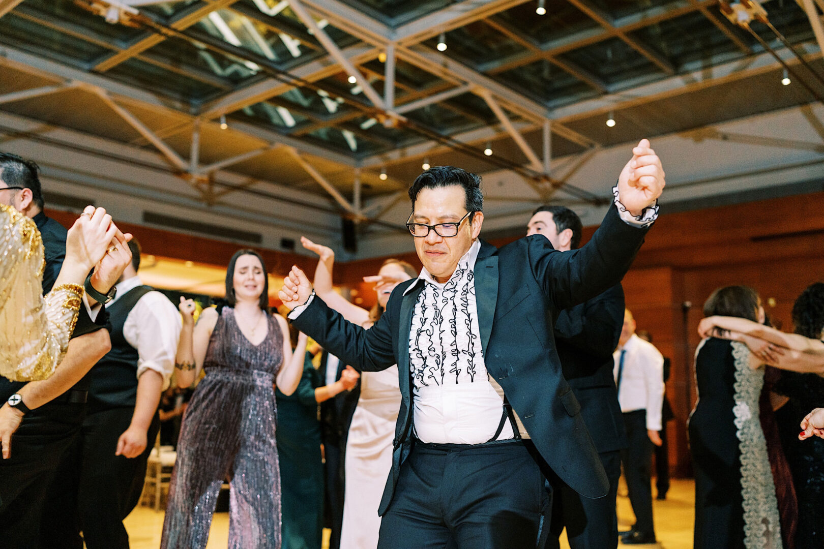 A man in a suit and bow tie dances energetically at a New Year's Eve event at the Kimmel Center in Philadelphia, surrounded by other people dancing in a well-lit hall with a high ceiling.