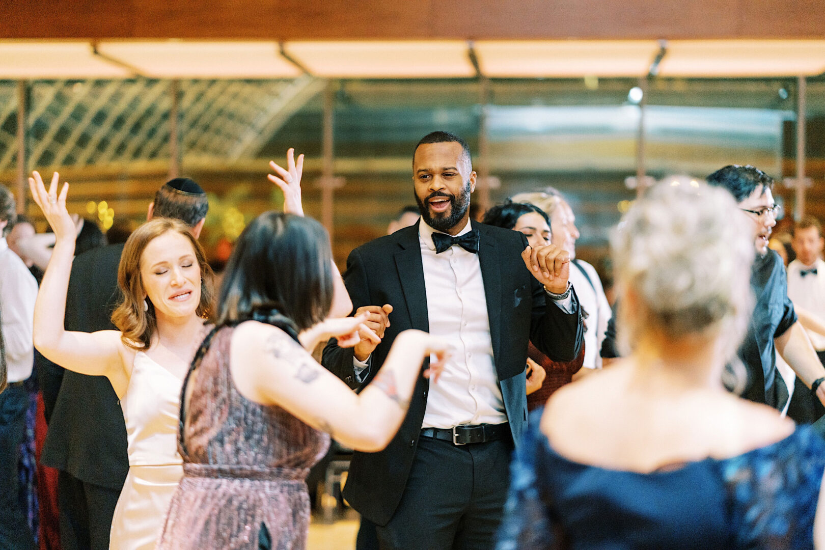 A group of people in formal attire dance enthusiastically at an indoor New Year's Eve event with large glass windows in the background, possibly at Kimmel Center in Philadelphia.