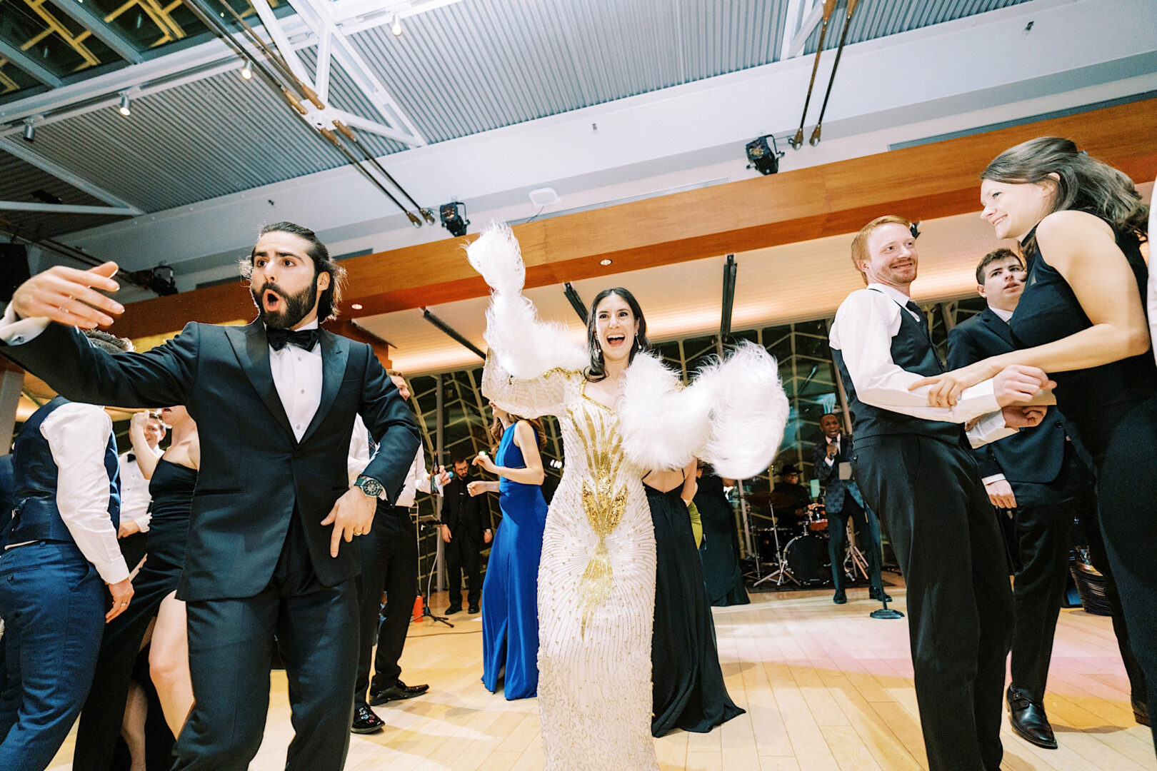 A group of people in formal attire dancing energetically at an indoor wedding, with a woman in a white gown and feathered sleeves in the center raising her arms.