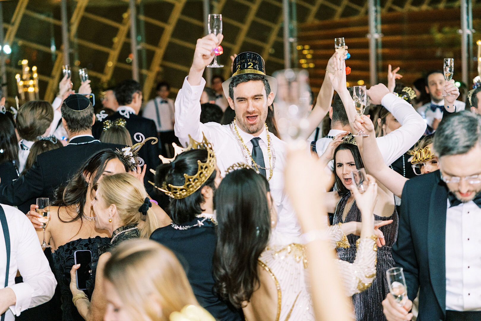 A group of people celebrating New Year's Eve at the Kimmel Center in Philadelphia, wearing party hats and crowns, raising champagne glasses in a crowded, festive atmosphere.