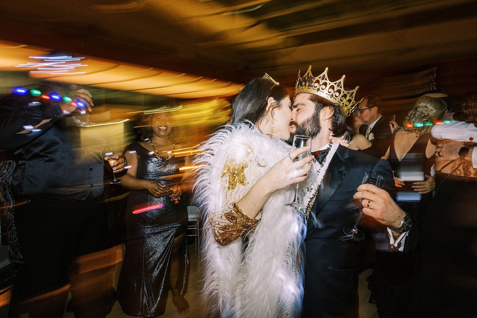 Two people wearing crowns and elegant attire share a close moment while holding champagne glasses at a lively New Year's Eve party. Blurred motion effects suggest dancing and movement in the background, capturing the festive spirit of Philadelphia as they celebrate together.