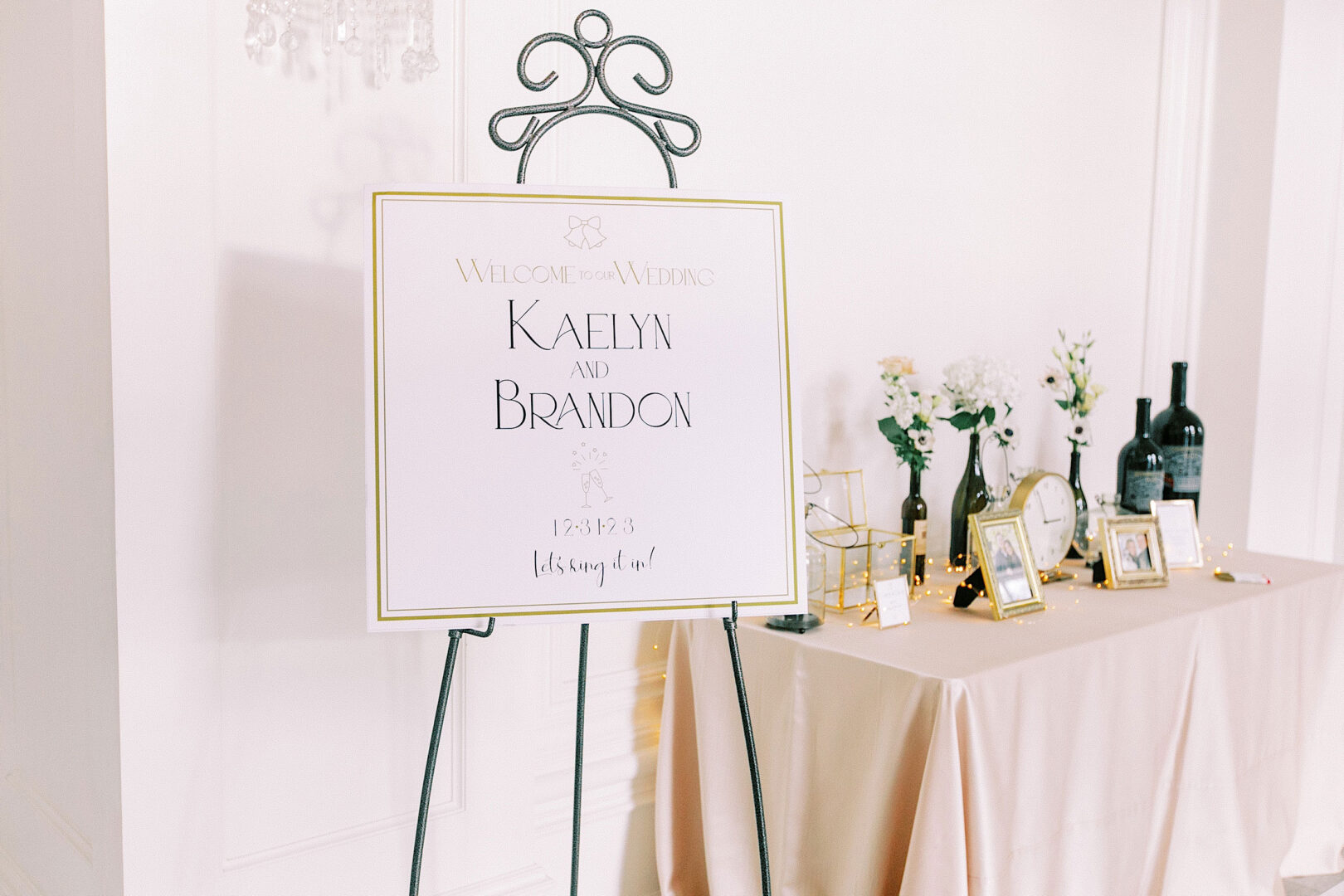 Welcome to the Ballroom at Ellis Preserve Wedding. Next to you is a charming welcome sign set by a table adorned with framed photos, lovely flowers, and decorative bottles, creating a picturesque start to this special day.