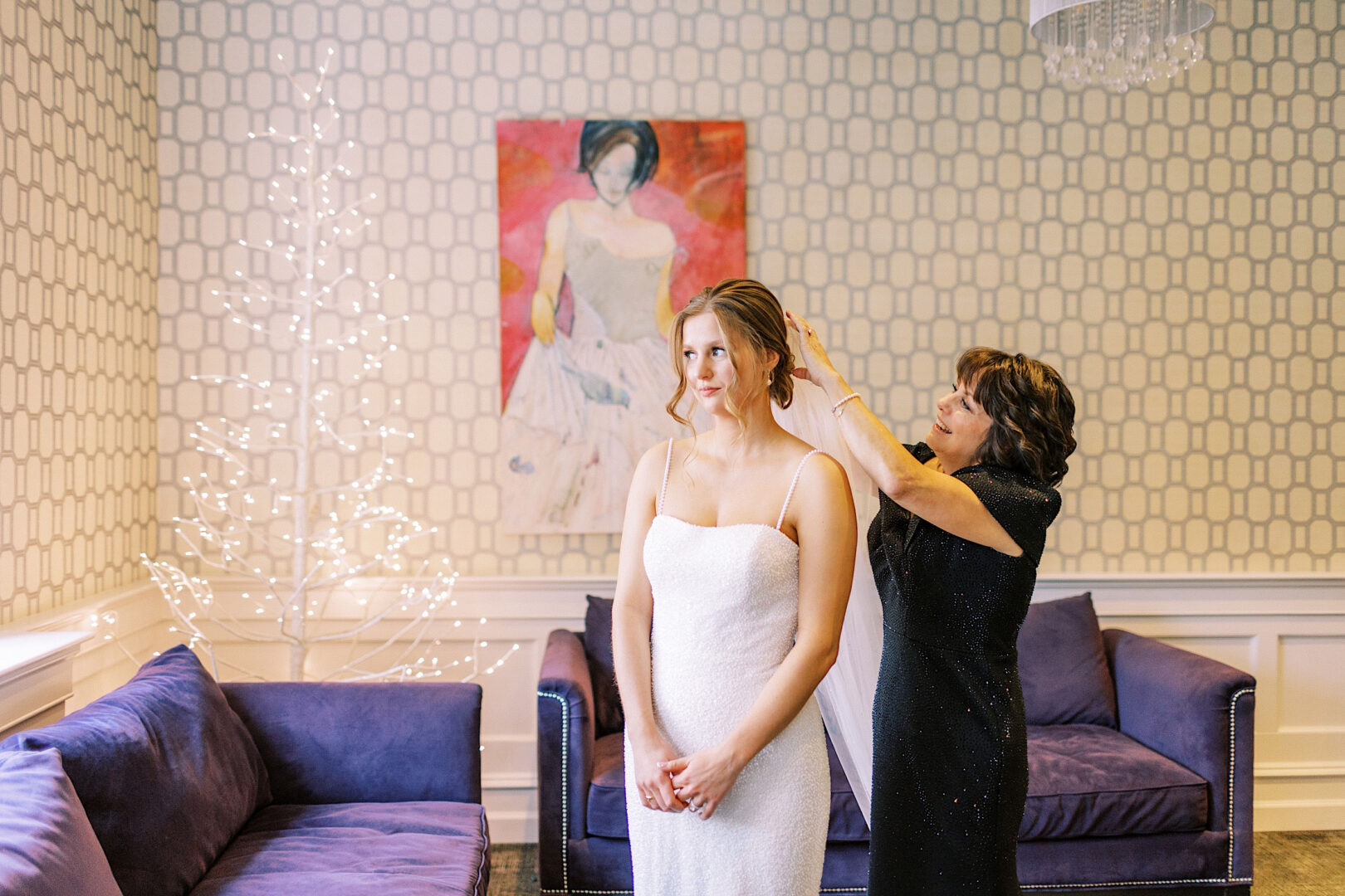 In the elegant Ballroom at Ellis Preserve, a woman gracefully adjusts the veil of a bride in a white dress, surrounded by a beautifully decorated room with purple couches and a charming painting on the wall.