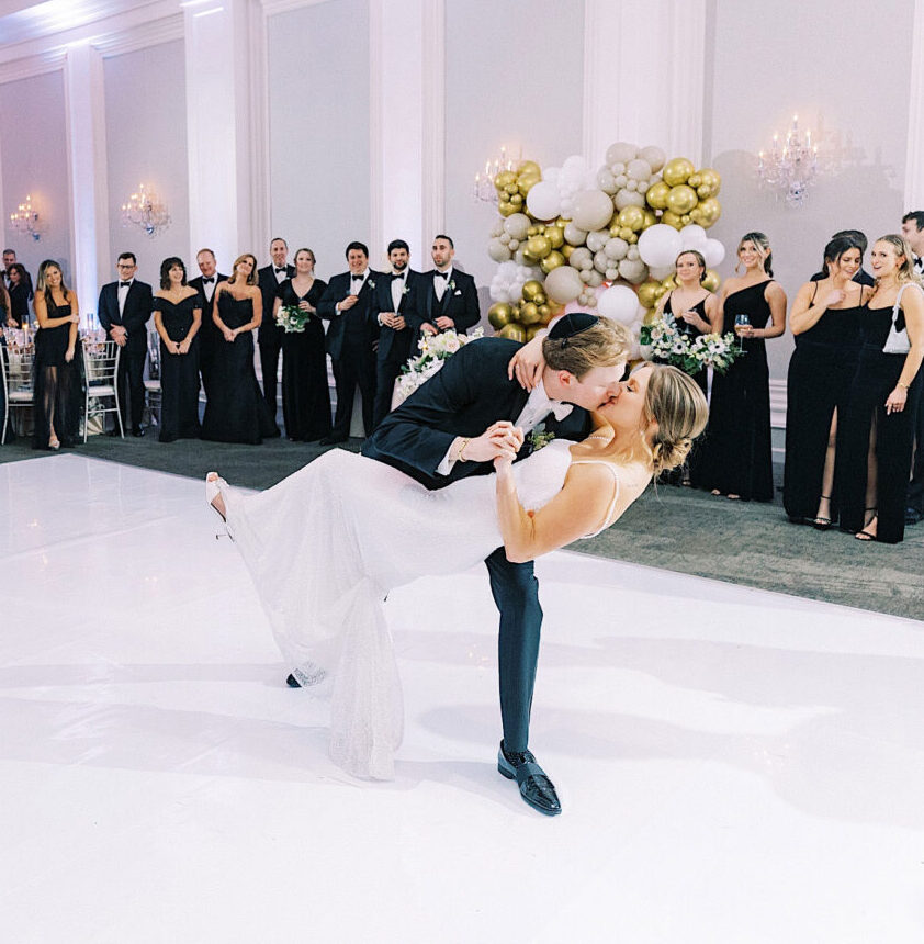 At a Ballroom at Ellis Preserve Wedding, a couple shares a dramatic dance dip on the pristine white floor, surrounded by guests and elegantly decorated with balloons.