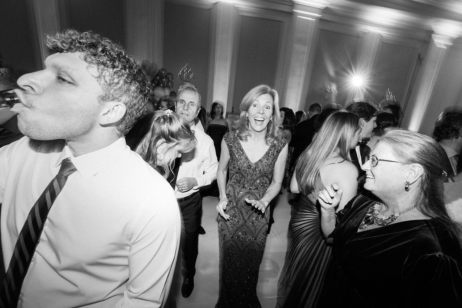 A man drinks from a bottle while a woman in an evening gown smiles widely, capturing the elegance of a Ballroom at Ellis Preserve wedding. Other guests gather in the dimly lit room, immersed in celebration.
