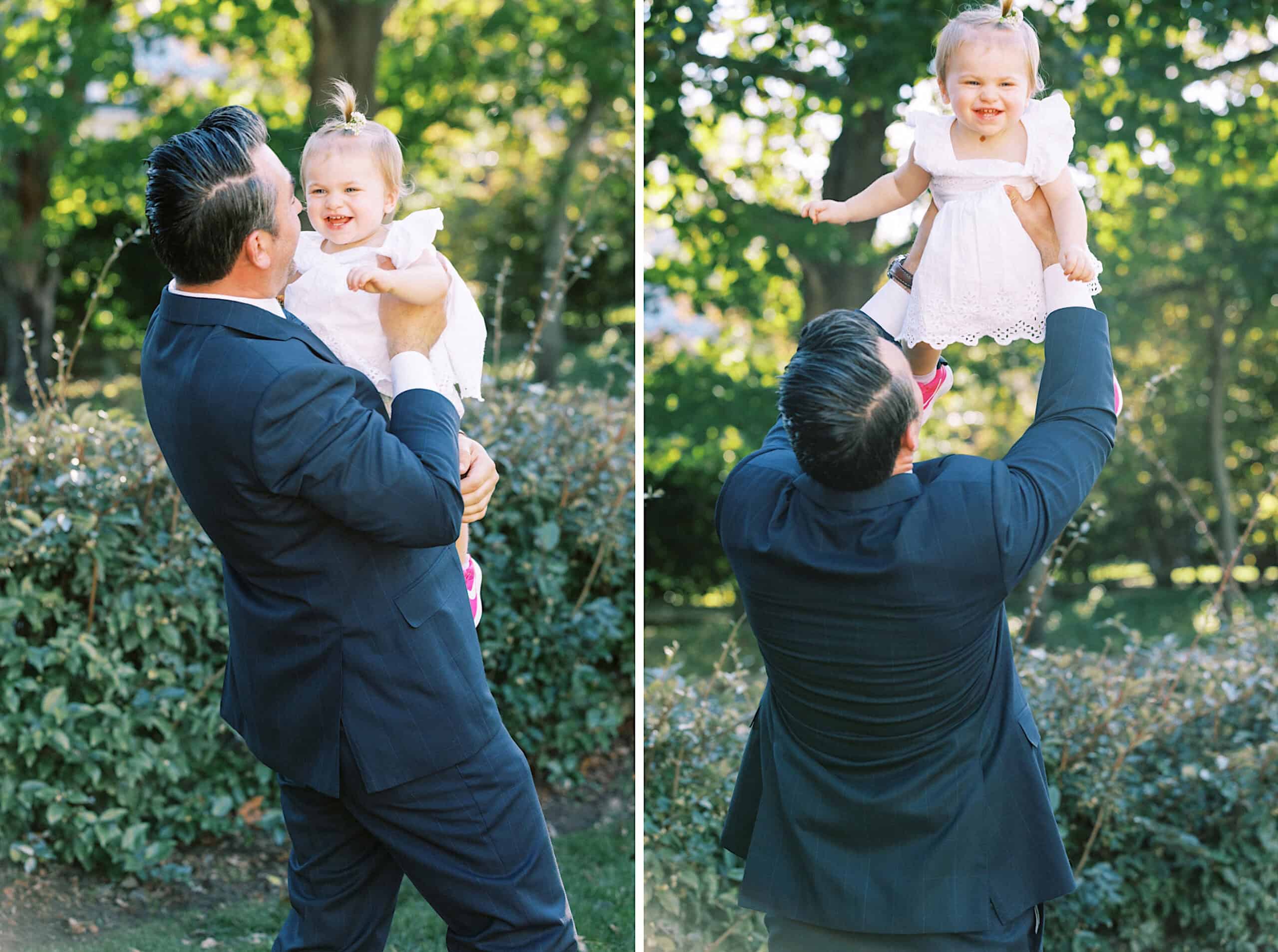 A man in a dark suit lifts a smiling toddler in a white dress outdoors at a Beach Plum Farm Wedding. They are surrounded by greenery and dappled sunlight.