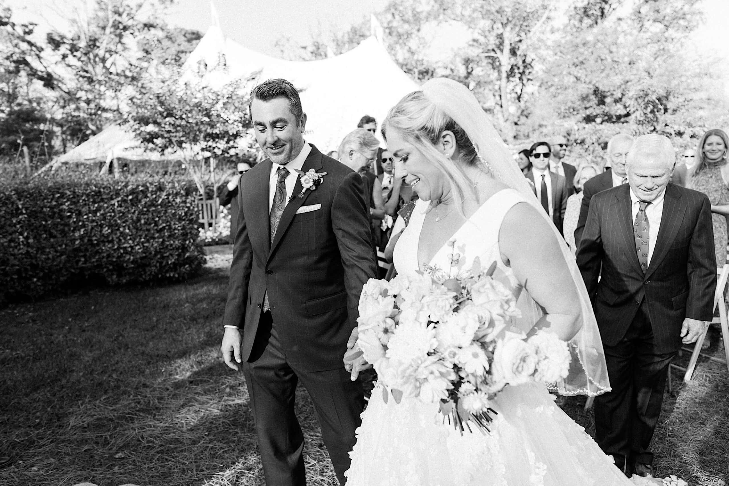 A bride and groom walk outdoors, holding hands and smiling, followed by guests at their Beach Plum Farm Wedding ceremony.