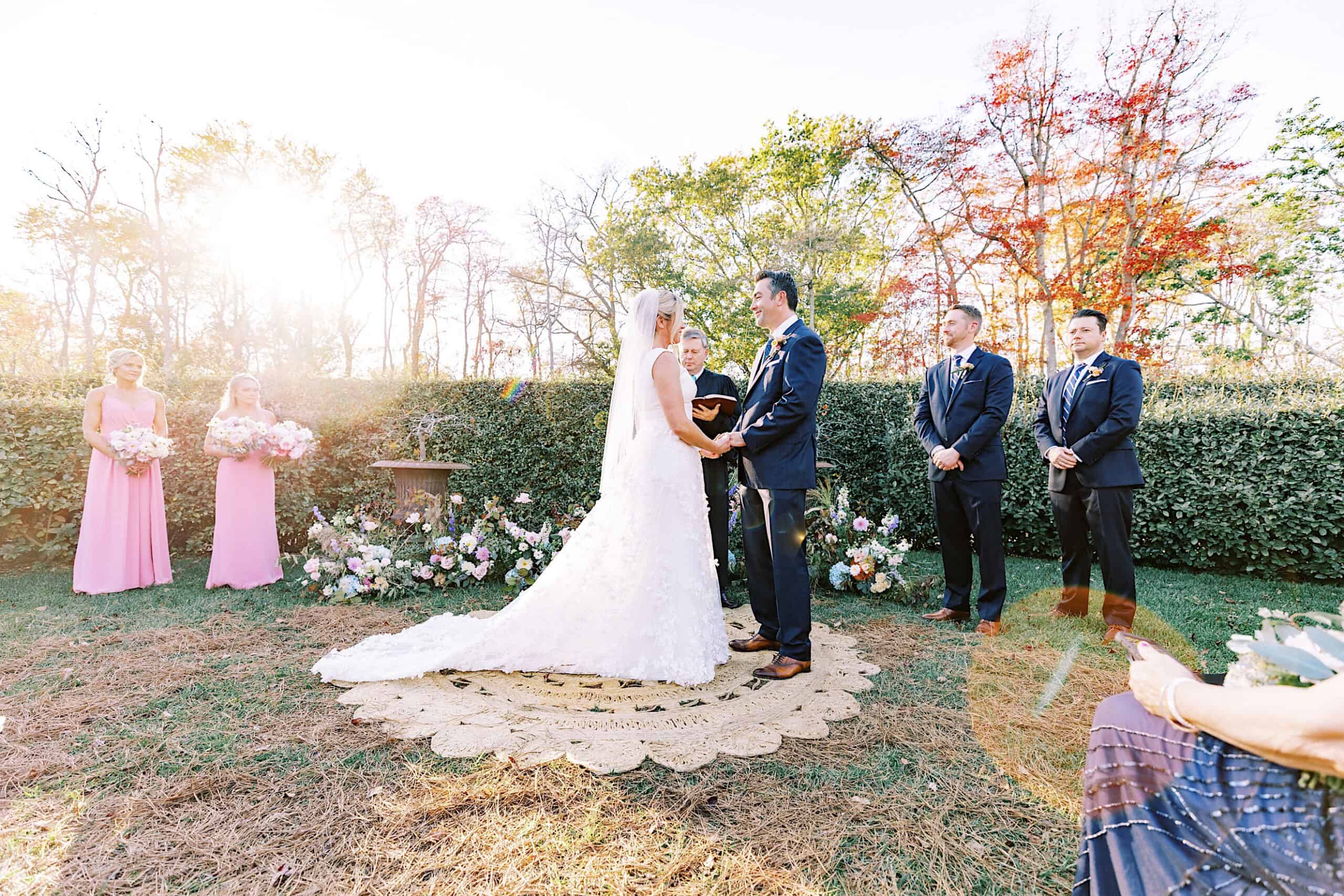A bride and groom stand facing each other holding hands during an outdoor Beach Plum Farm Wedding ceremony, with bridesmaids and groomsmen beside them.