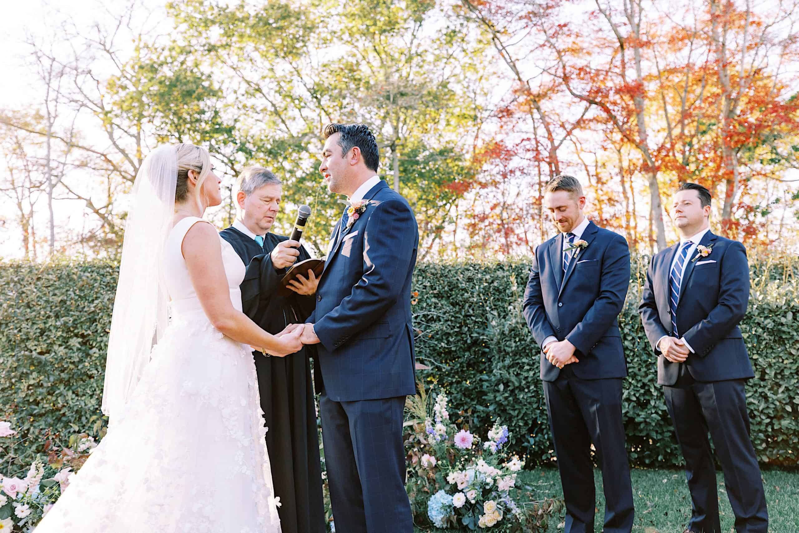 A bride and groom stand facing each other holding hands during an outdoor Beach Plum Farm Wedding ceremony, with an officiant and two groomsmen present.