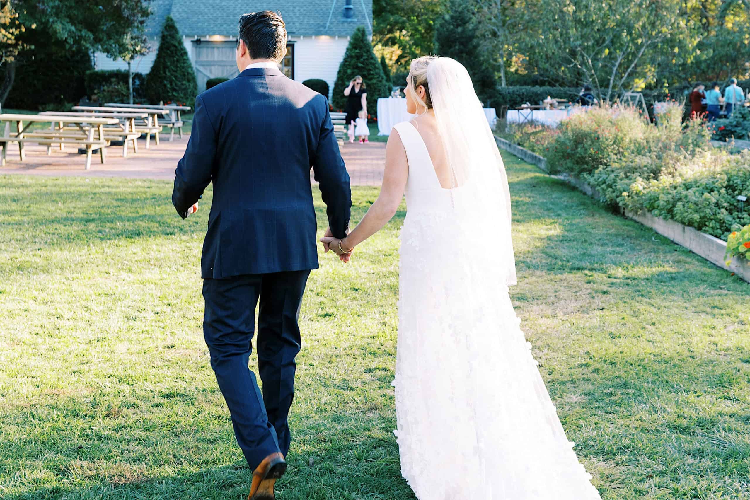 A bride and groom walk hand in hand on grass at an outdoor Beach Plum Farm Wedding venue, with gardens and picnic tables visible in the background.