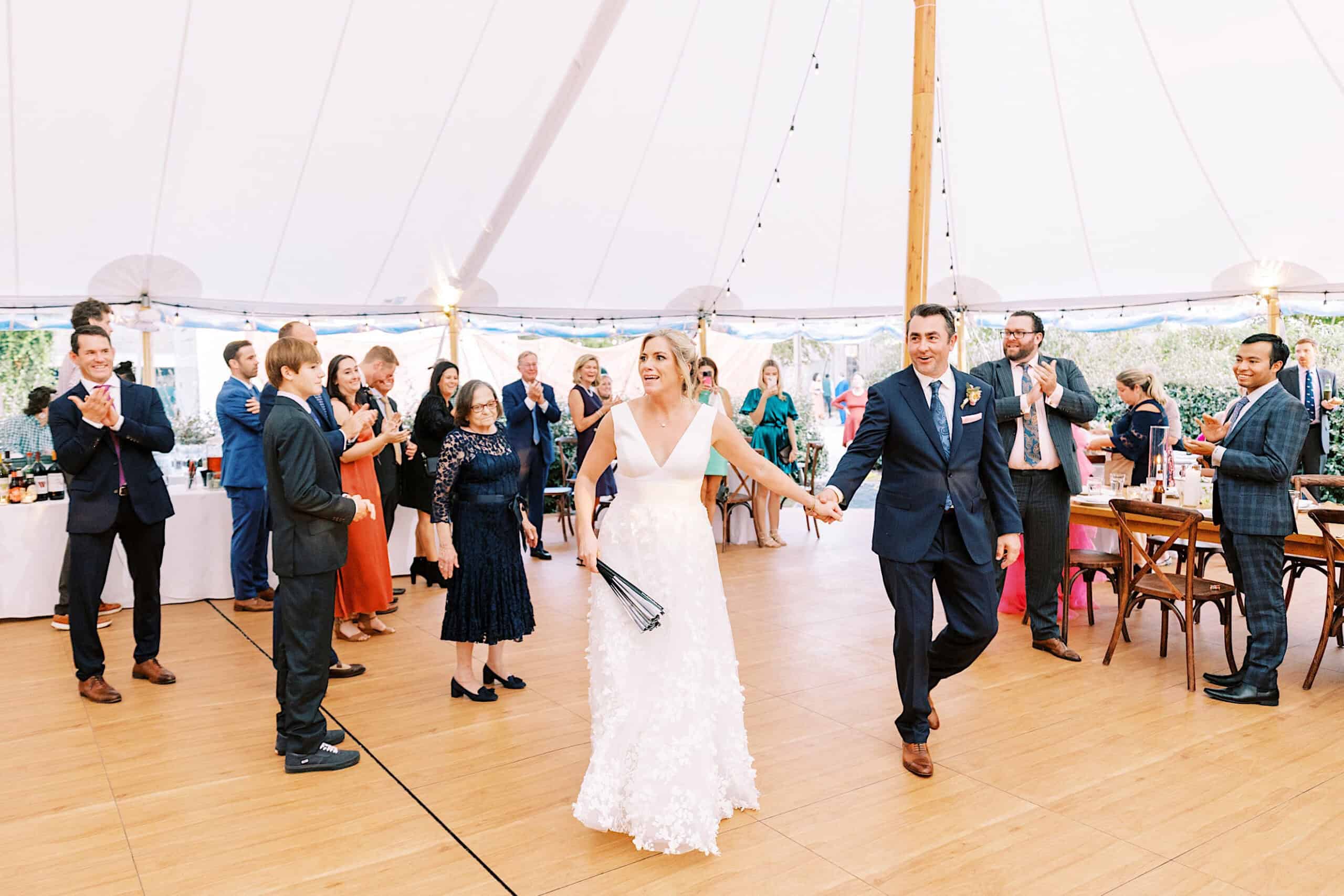 A bride and groom hold hands and enter a decorated tent at their Beach Plum Farm Wedding while guests stand and clap, some smiling and others taking photos.