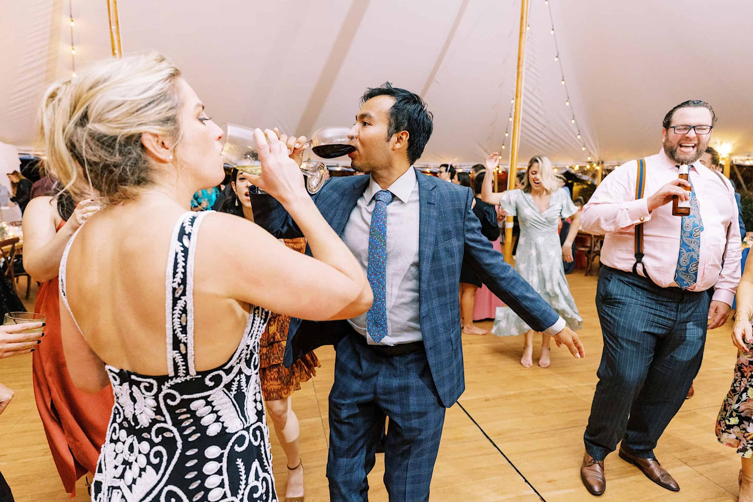 Guests in formal attire dance and drink at an indoor Beach Plum Farm Wedding, celebrating under a decorated tent adorned with glowing string lights.