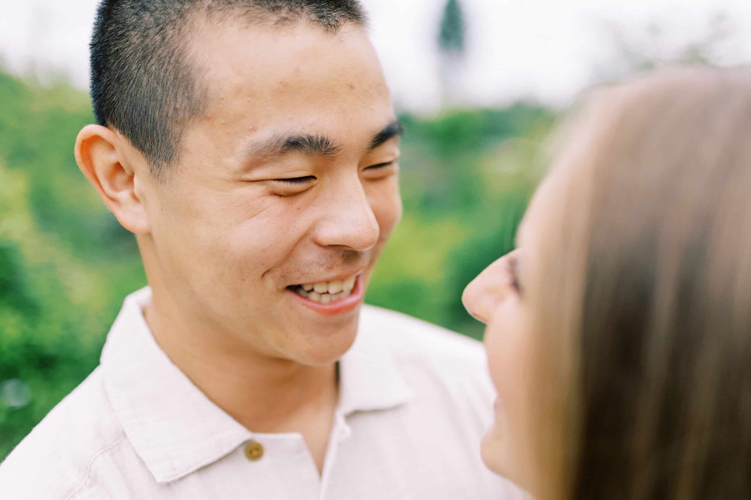 A man smiles and looks at a woman outdoors, with greenery blurred in the background during their Engagement Photo Session at Twinning Valley Park.