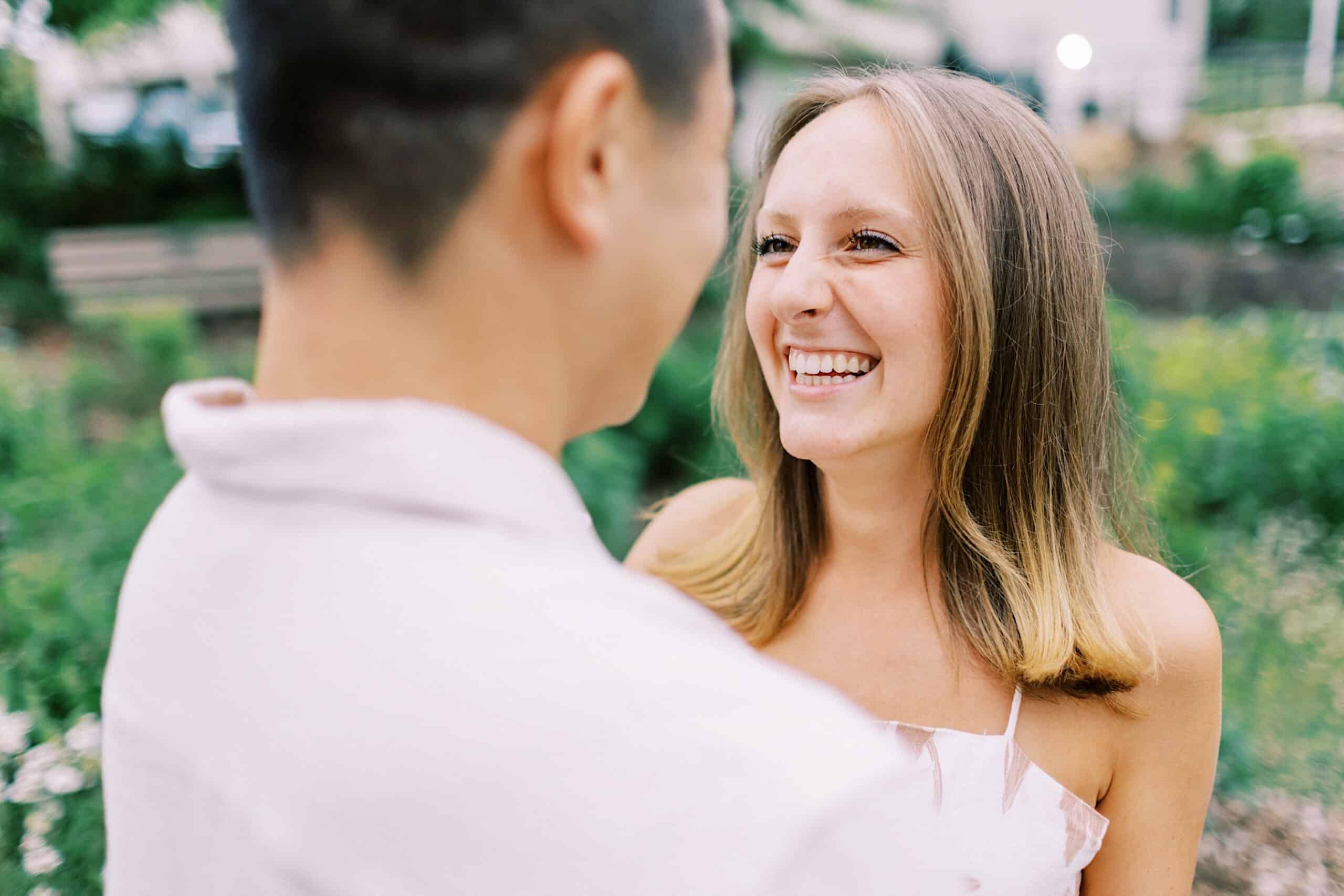 A woman smiles joyfully at a man outdoors in a garden, capturing the happiness of their Engagement Photo Session at Twinning Valley Park. The focus is on her radiant expression amidst the scenic park setting.