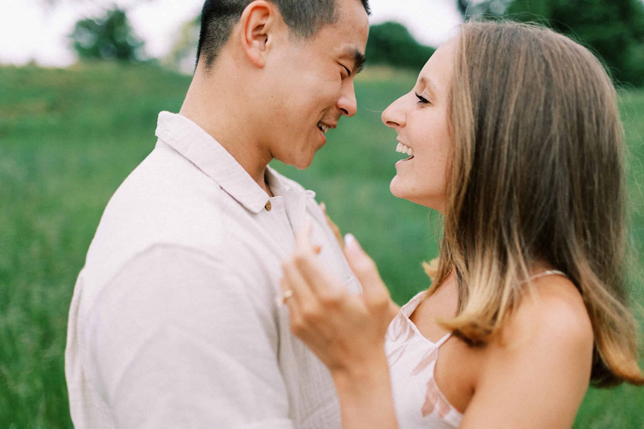 A man and a woman stand close together outdoors, smiling at each other in a grassy area with trees in the background during their engagement photo session at Twinning Valley Park.