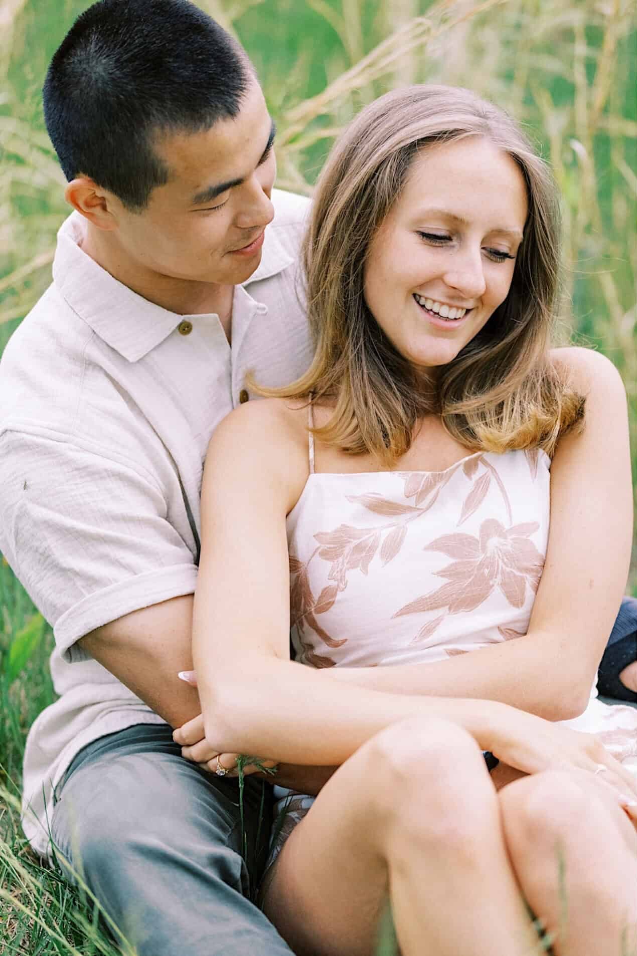 A couple sits together on the grass, embracing and smiling at each other during an engagement photo session at Twinning Valley Park, surrounded by a beautiful natural outdoor setting.