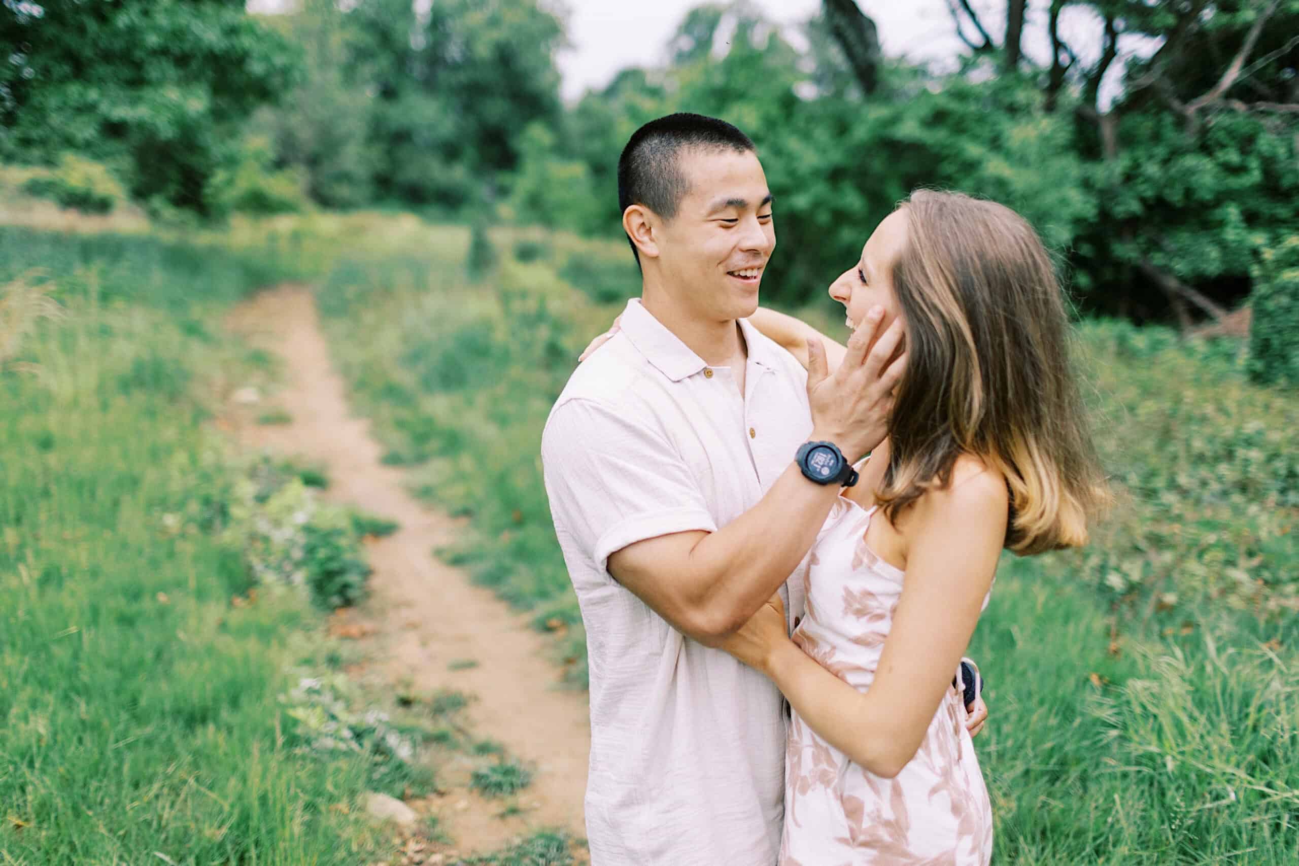 A man and woman stand on a dirt path in a green, wooded area during their Engagement Photo Session at Twinning Valley Park. The man is touching the woman’s face, and both are smiling at each other.