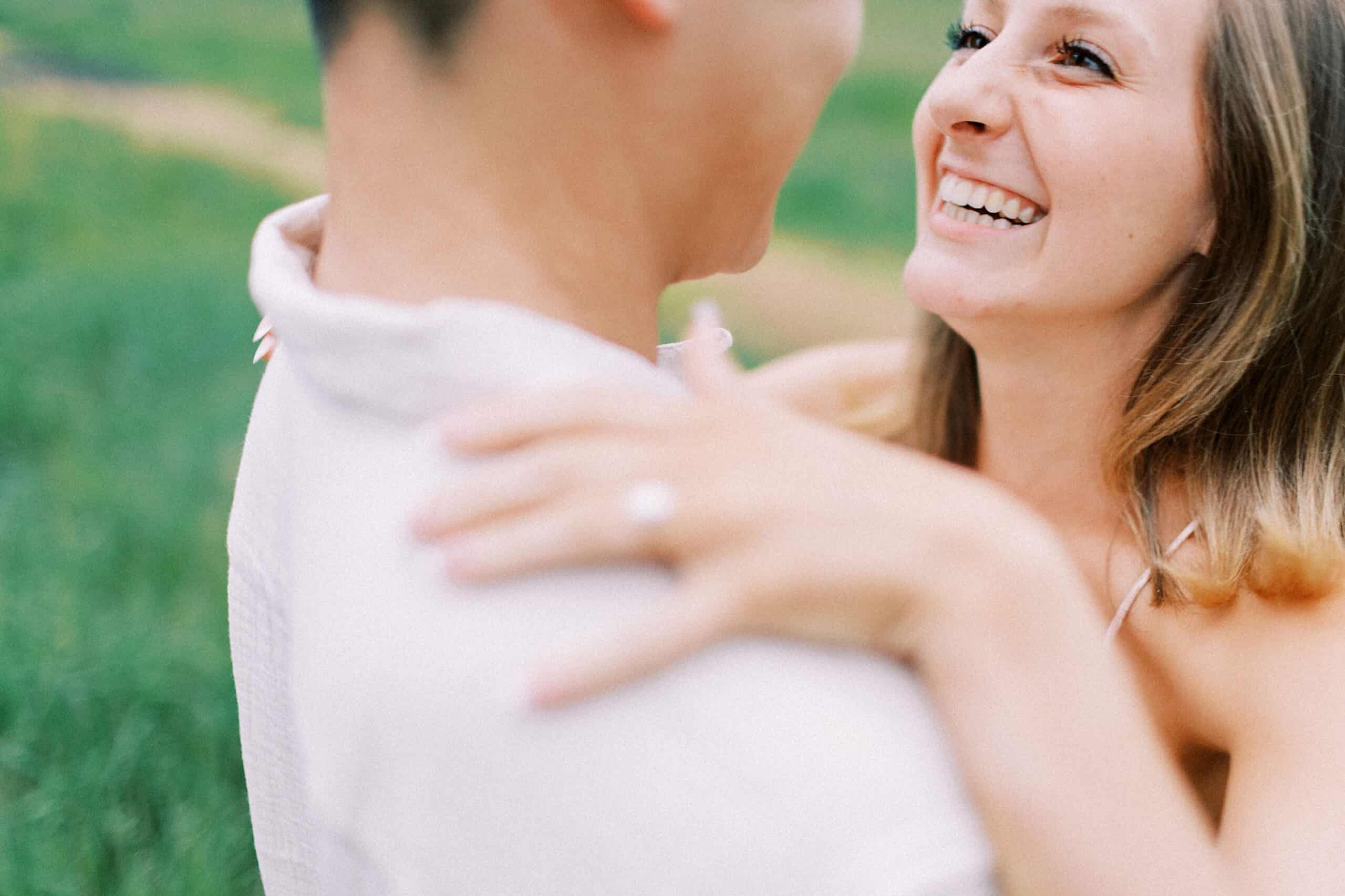 A woman smiles while embracing a man outdoors at their Engagement Photo Session at Twinning Valley Park, her hand resting on his shoulder with a ring visible on her finger. The grassy background is softly blurred.