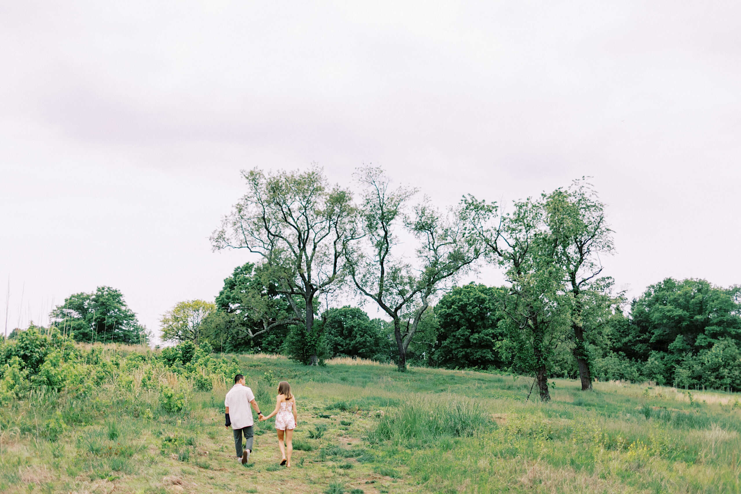 Two people walk together through a grassy field with scattered trees under a cloudy sky during their engagement photo session at Twinning Valley Park.