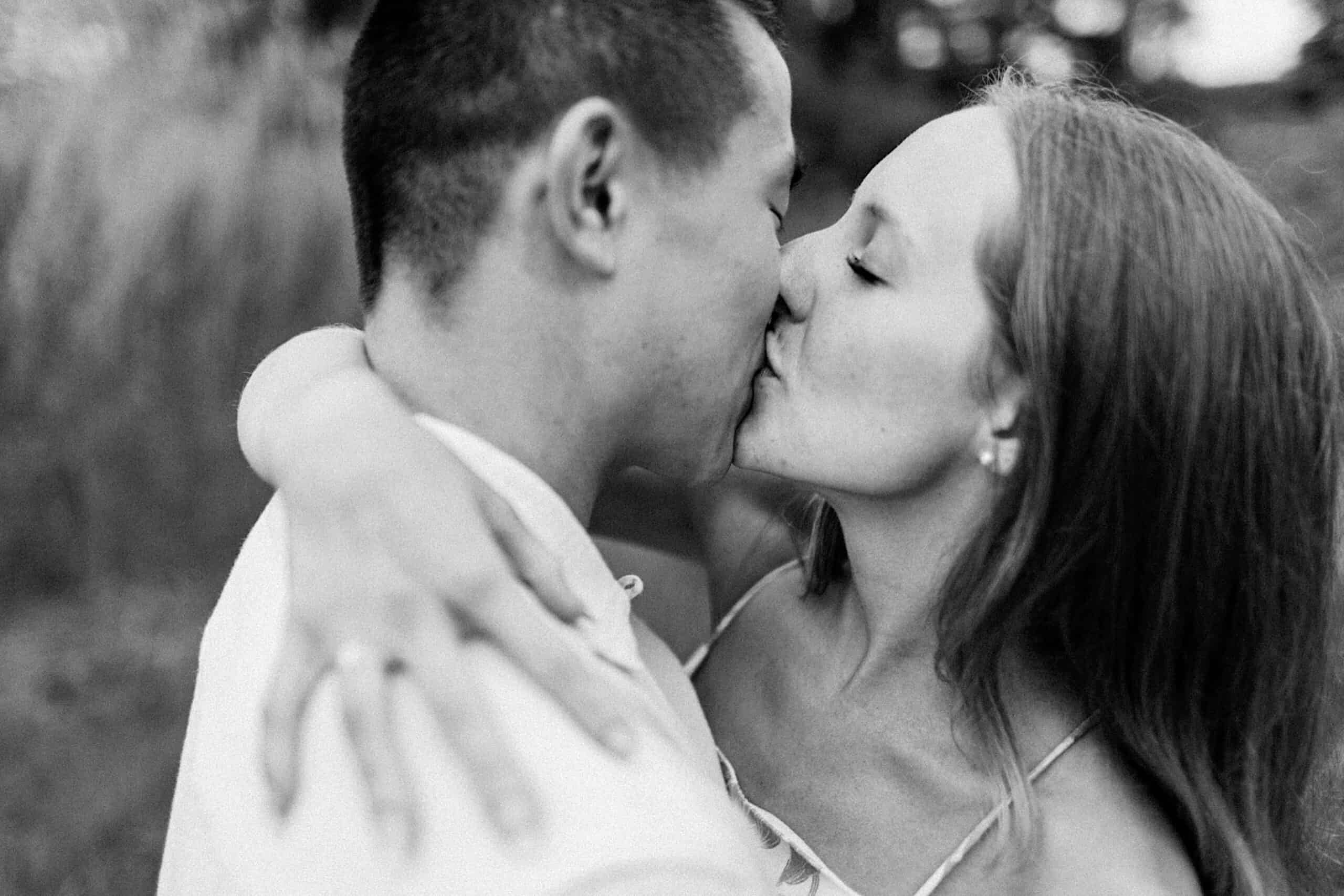 A close-up, black-and-white photo of a couple kissing outdoors during their engagement photo session at Twinning Valley Park, with the woman's arms wrapped around the man's shoulders.