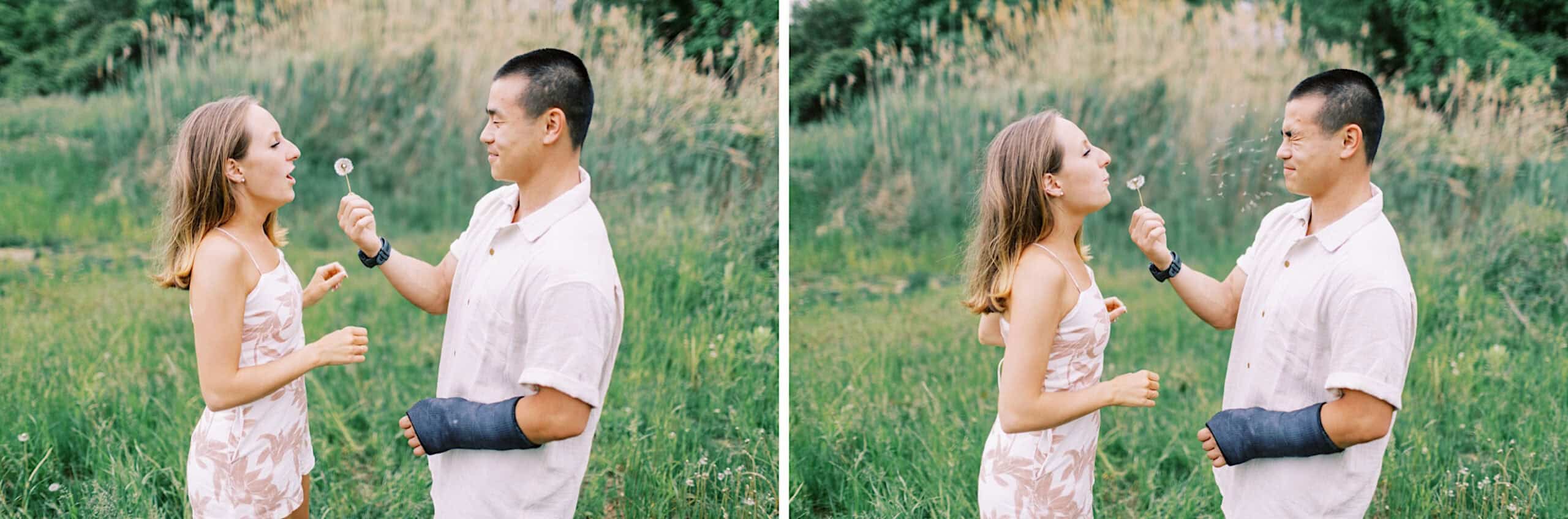Two people stand in a grassy field at Twinning Valley Park; one holds a dandelion while the other blows on it, scattering seeds in the air during their engagement photo session.
