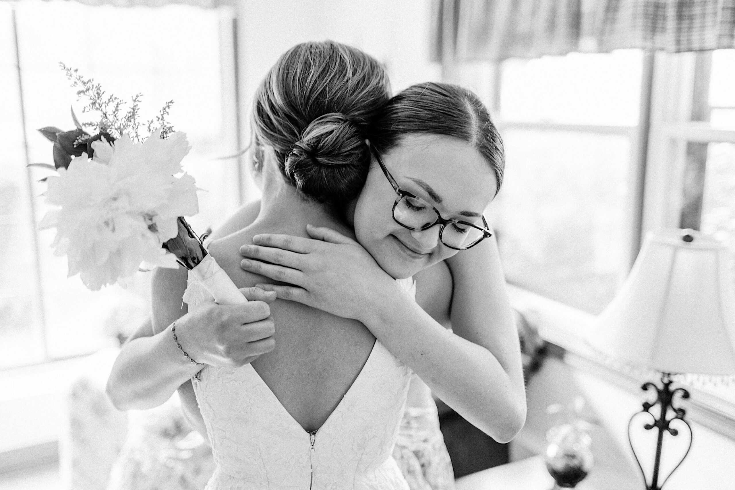 Two women embrace indoors; one is holding a bouquet and wearing a wedding dress, while the other, in glasses, smiles with her eyes closed—capturing pure joy at a Summer Wedding at The High Point.