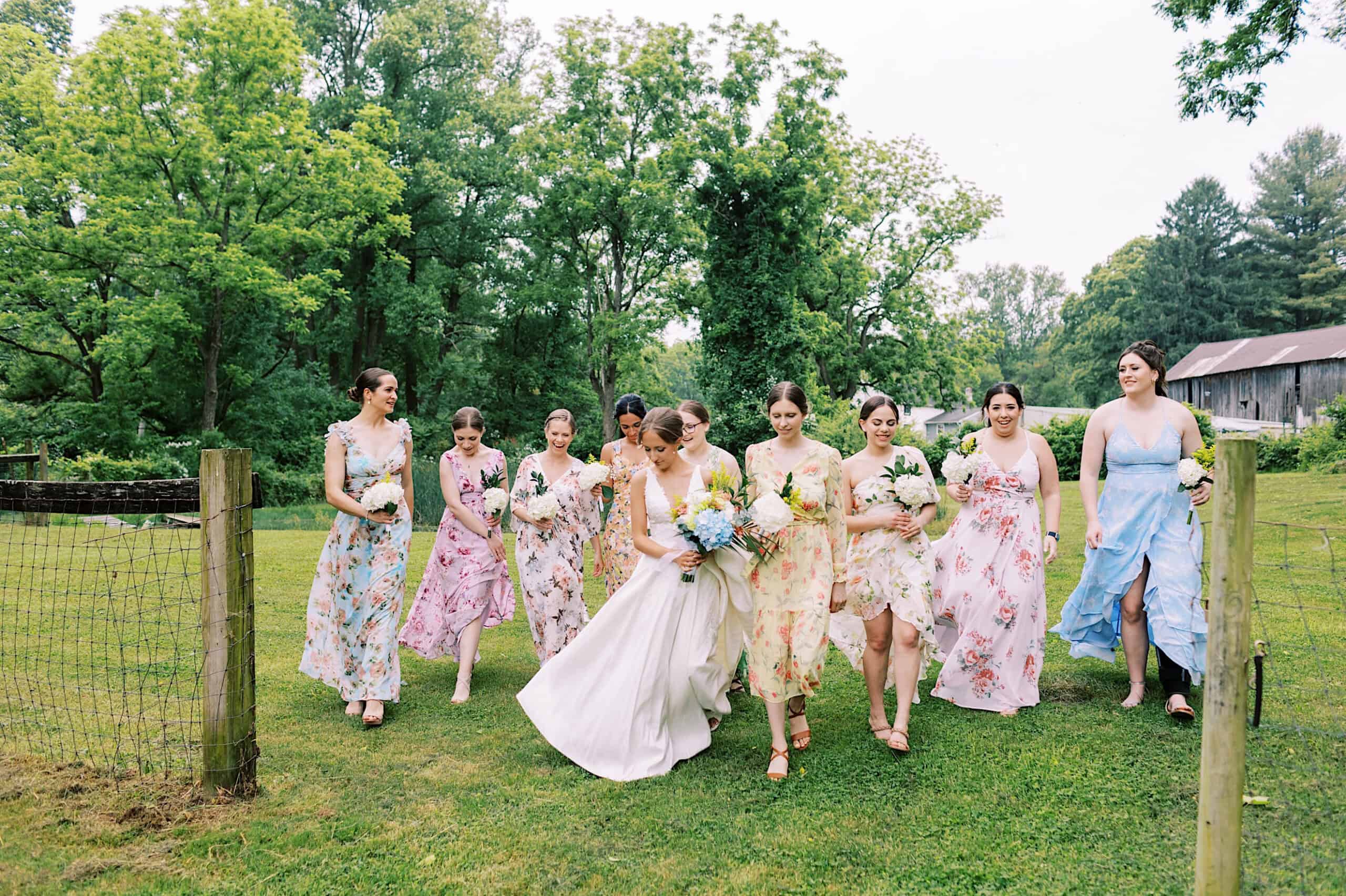 At a Summer Wedding at The High Point, a bride in white walks on grass with nine bridesmaids in floral dresses, all holding bouquets, surrounded by trees and a fence.