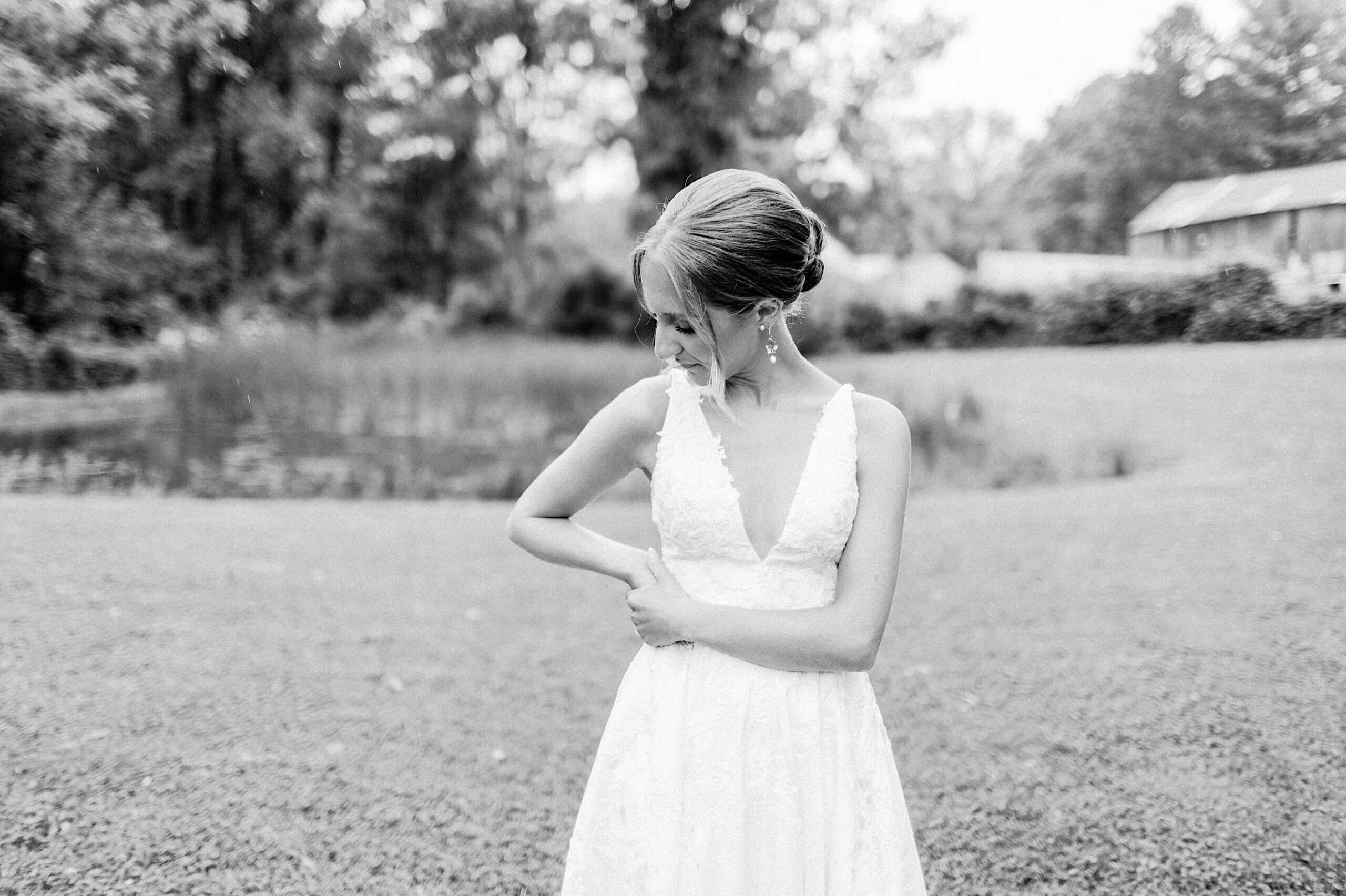 A woman in a sleeveless white dress stands outdoors on grass at a Summer Wedding at The High Point, looking down with one hand on her waist. Trees and a pond are visible in the background. The image is in black and white.