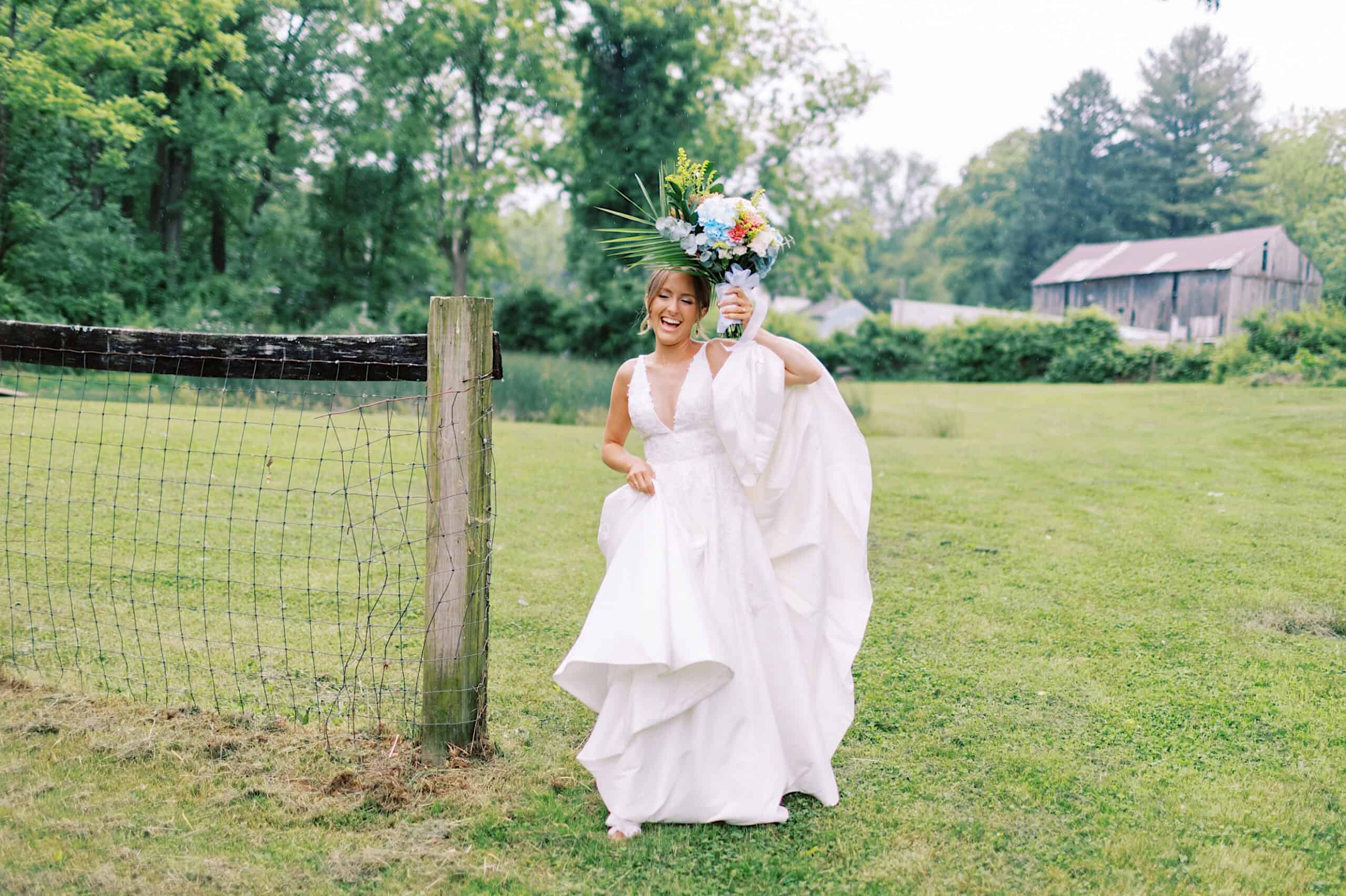 A bride in a white dress walks barefoot on grass, holding up her bouquet and her train, with a wire fence and barn in the background—capturing the magic of a Summer Wedding at The High Point.