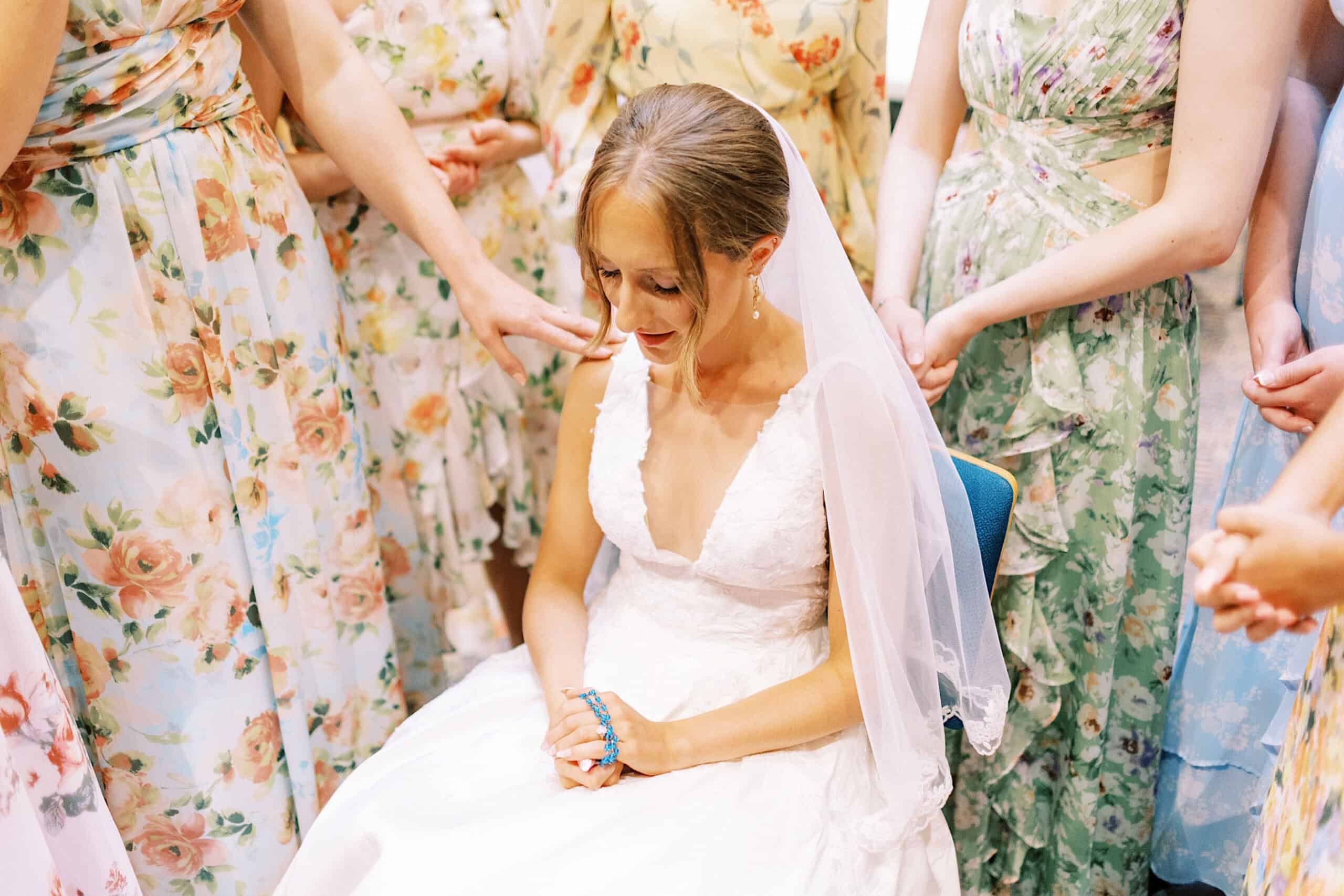 At a Summer Wedding at The High Point, a bride in a white dress and veil sits with her eyes closed, holding blue rosary beads, surrounded by women in floral dresses.