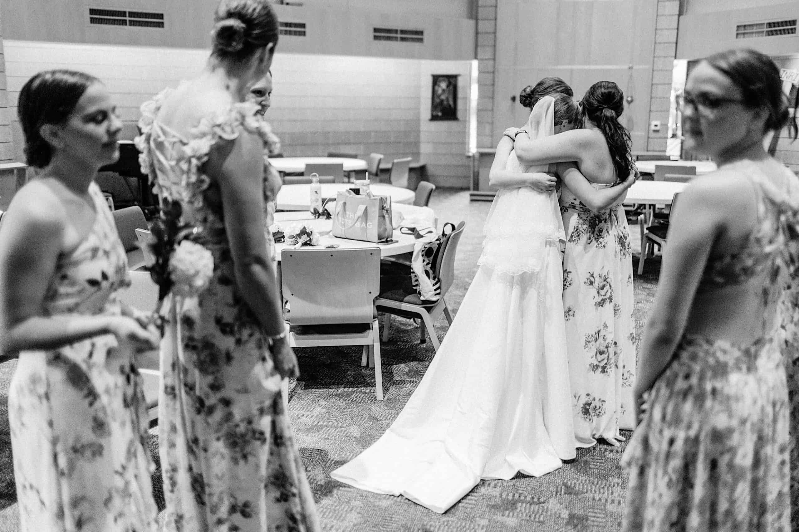 A bride in a wedding dress hugs a bridesmaid while other bridesmaids in floral dresses stand nearby in a room with tables and chairs during a Summer Wedding at The High Point.