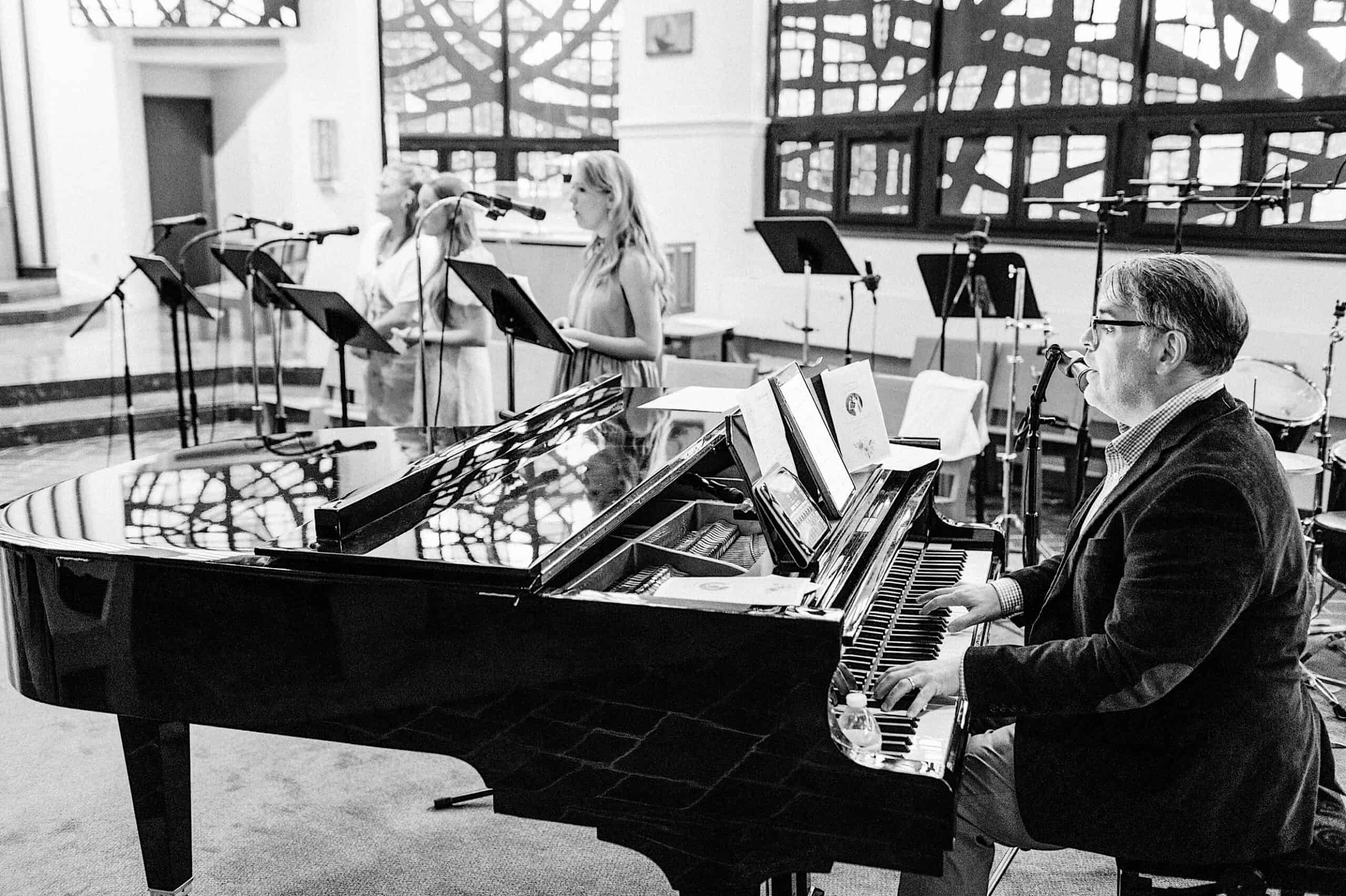 A man plays a grand piano while two women stand and sing at microphones in a room with large, geometric windows during a Summer Wedding at The High Point.