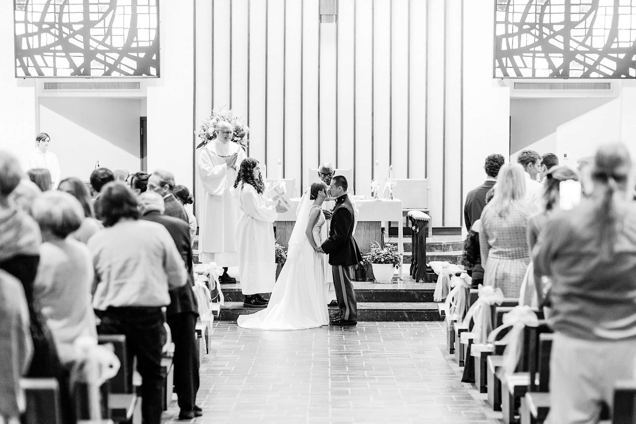 A bride and groom kiss at the altar during a Summer Wedding at The High Point, while guests stand in the pews and a priest looks on in the charming church.