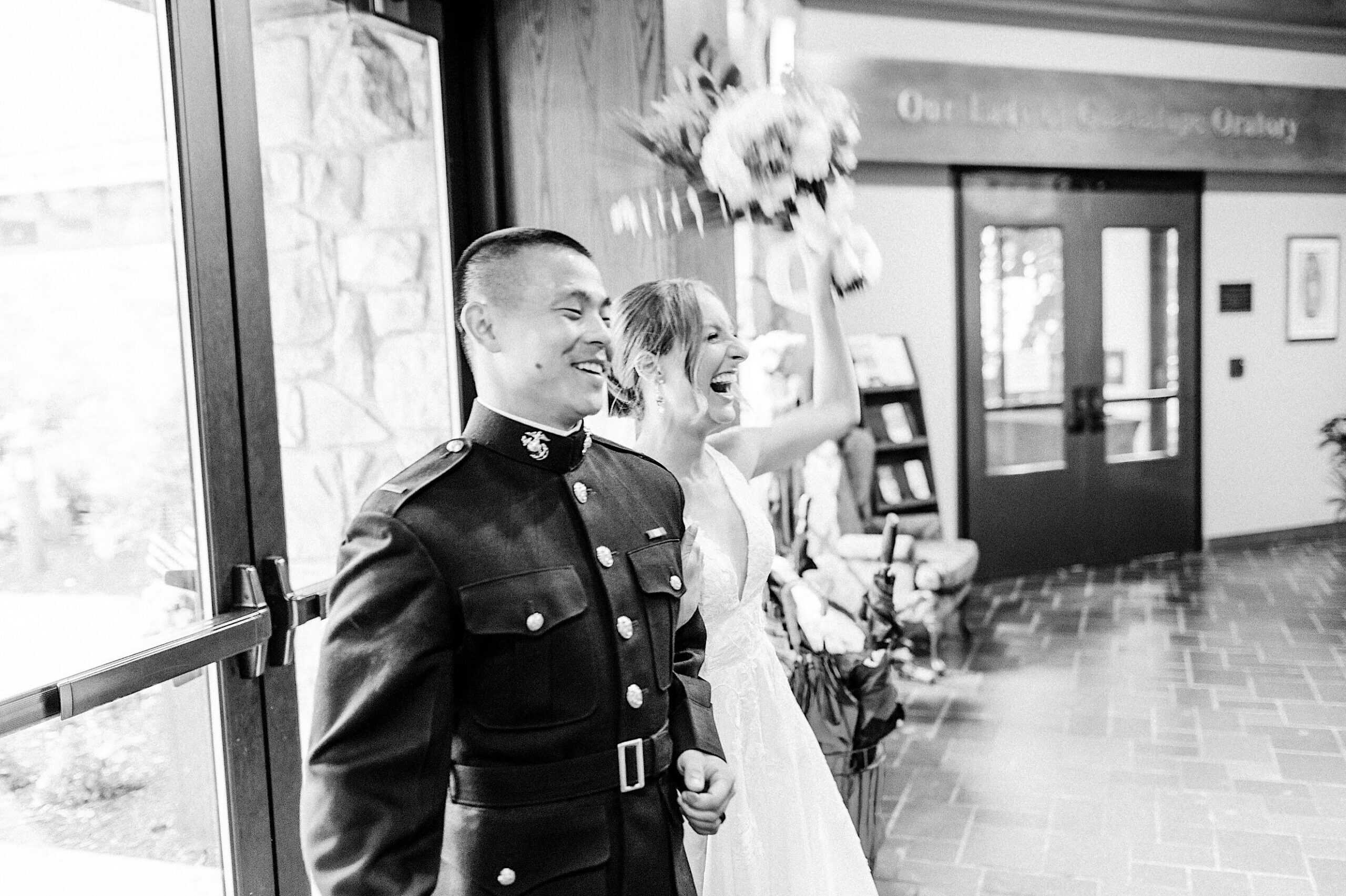 A man in a military uniform and a woman in a wedding dress stand together indoors, with the woman holding a bouquet and smiling, capturing a joyful moment from their Summer Wedding at The High Point.