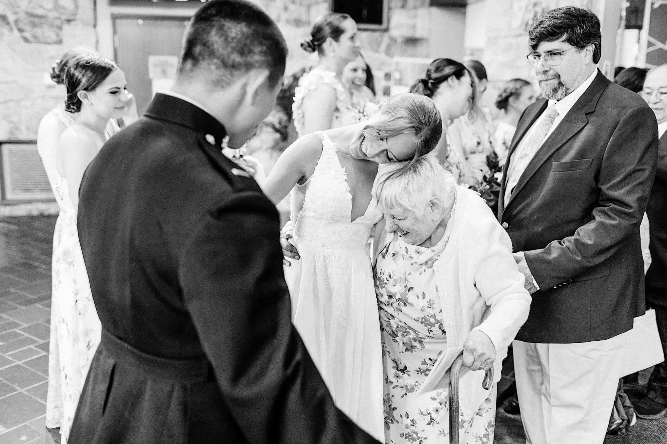 At a Summer Wedding at The High Point, a bride in a wedding dress hugs an elderly woman using a cane, surrounded by guests in formal attire at the gathering.