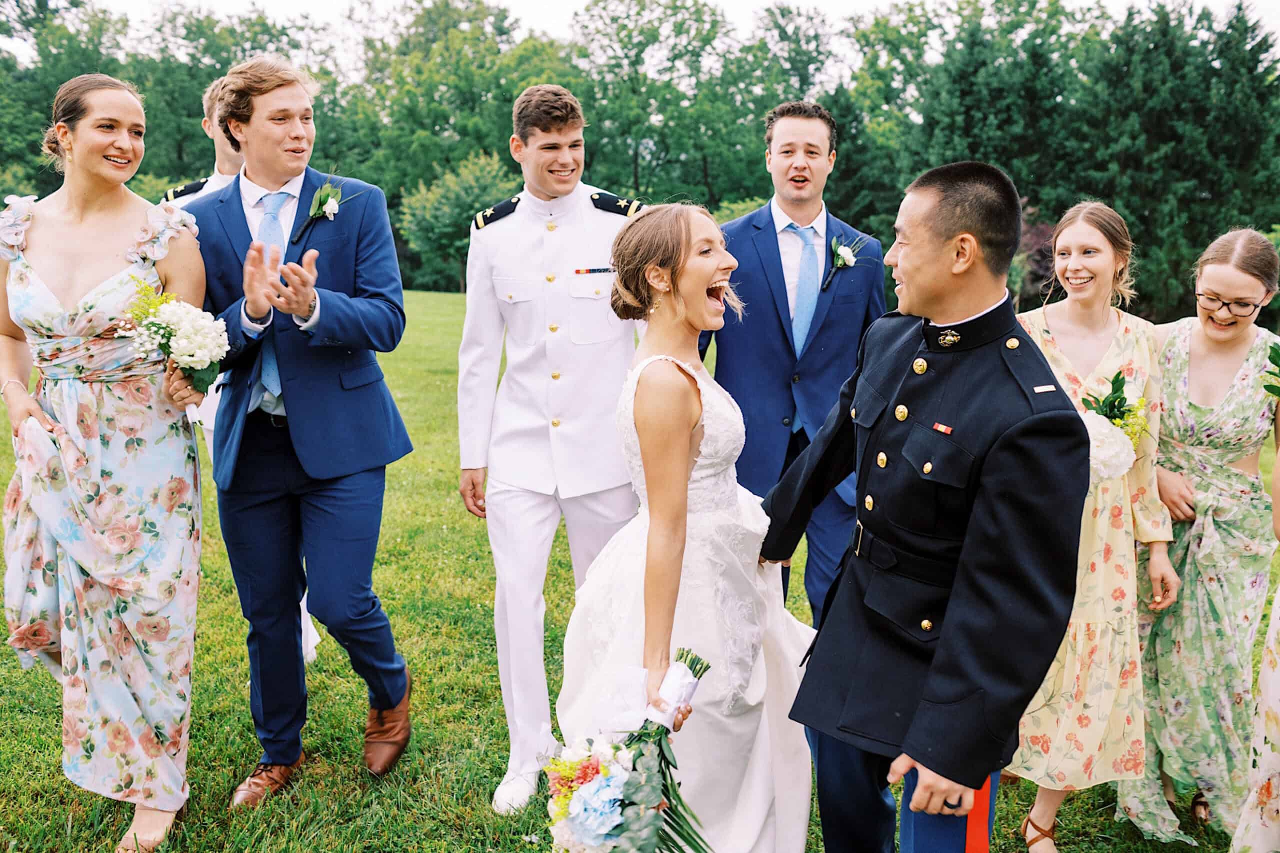 A bride and groom smile at each other while walking outside with their wedding party, dressed in dresses, suits, and military uniforms during their Summer Wedding at The High Point.