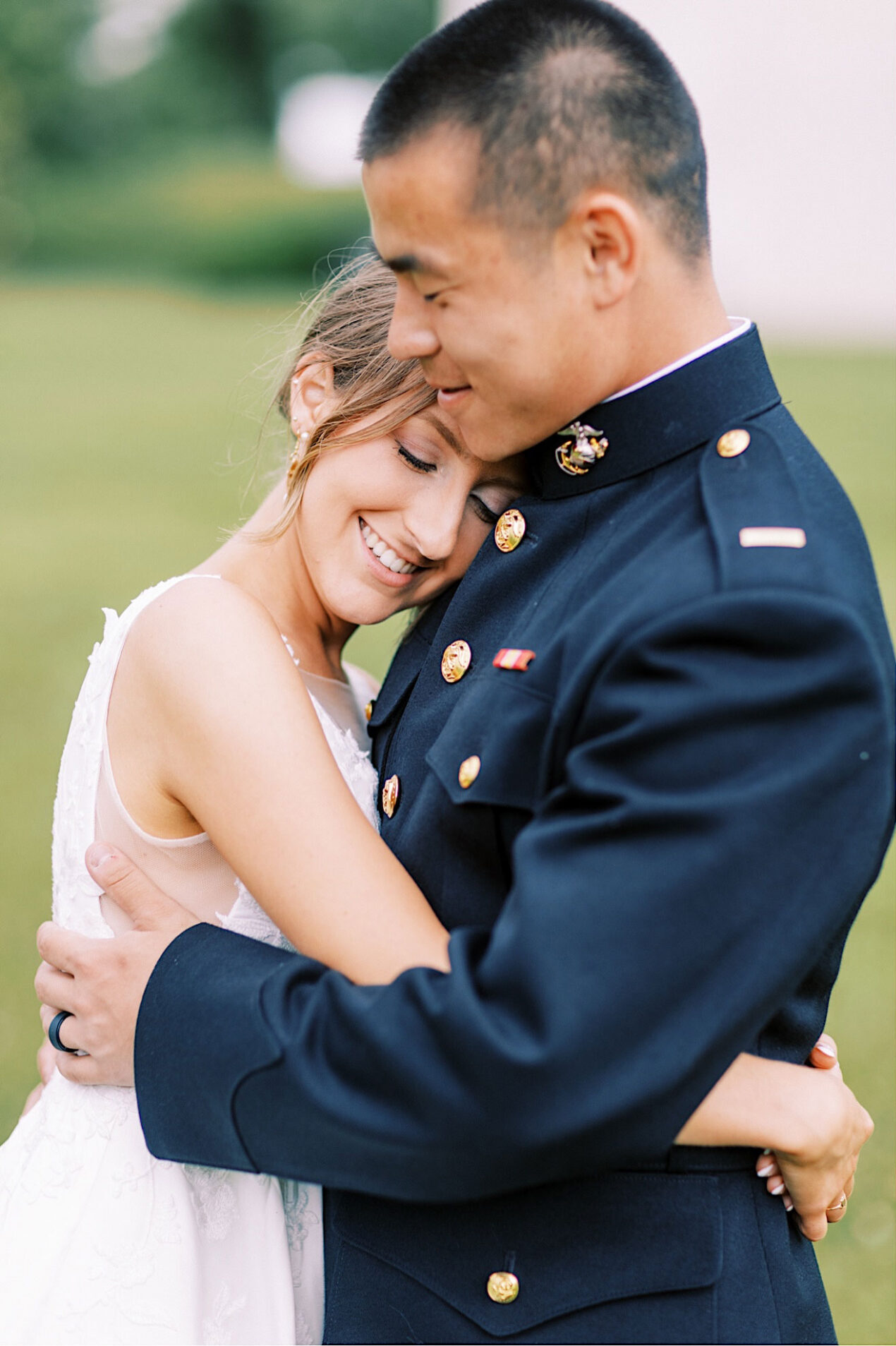 A couple embraces, with the man in a military uniform and the woman in a white dress—capturing the joy of a Summer Wedding at The High Point, as their bridal bouquet rests on lush green grass.