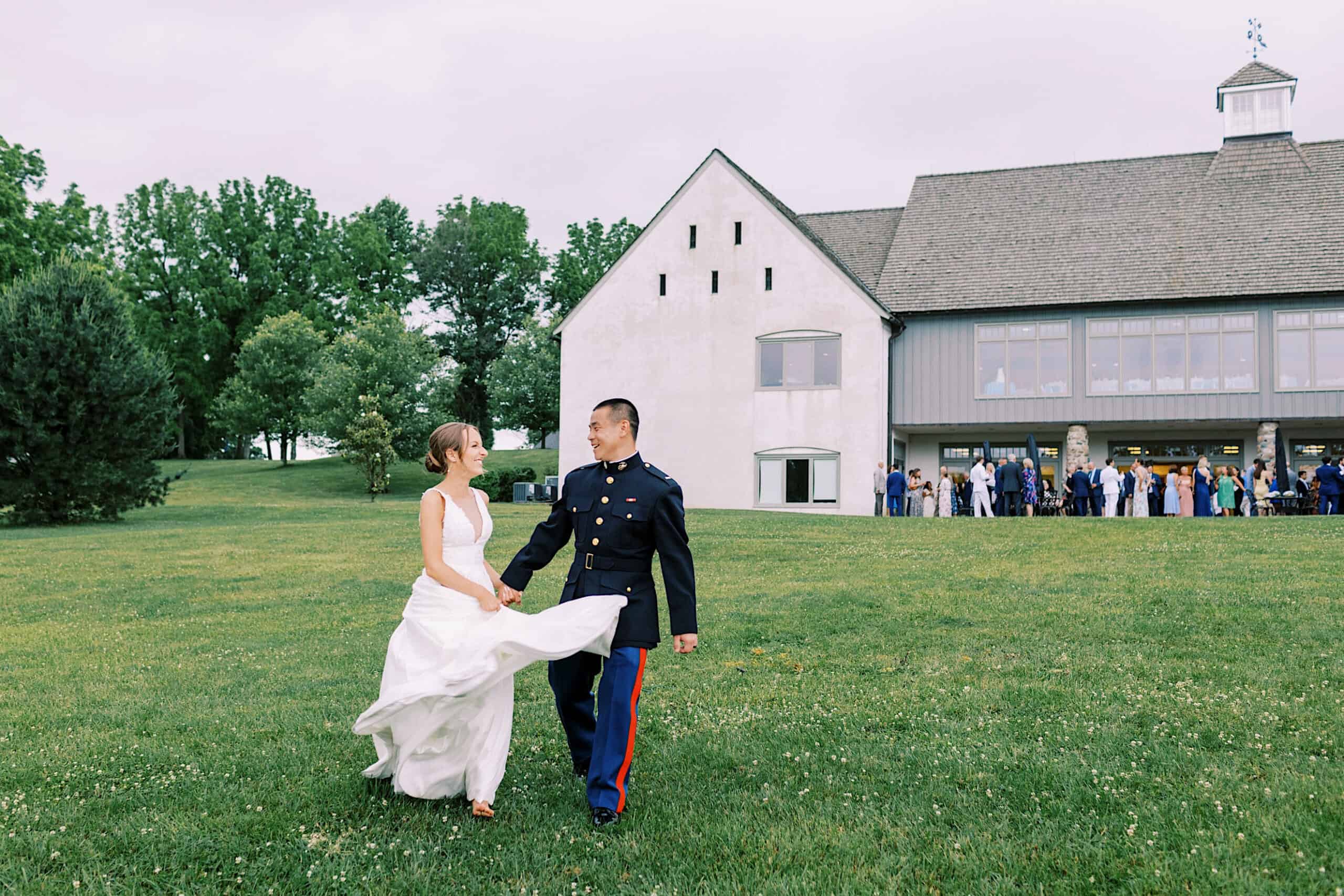 A bride in a white dress and a groom in a military uniform walk hand in hand on a grassy lawn during their Summer Wedding at The High Point, with guests gathered outside the large white building in the background.