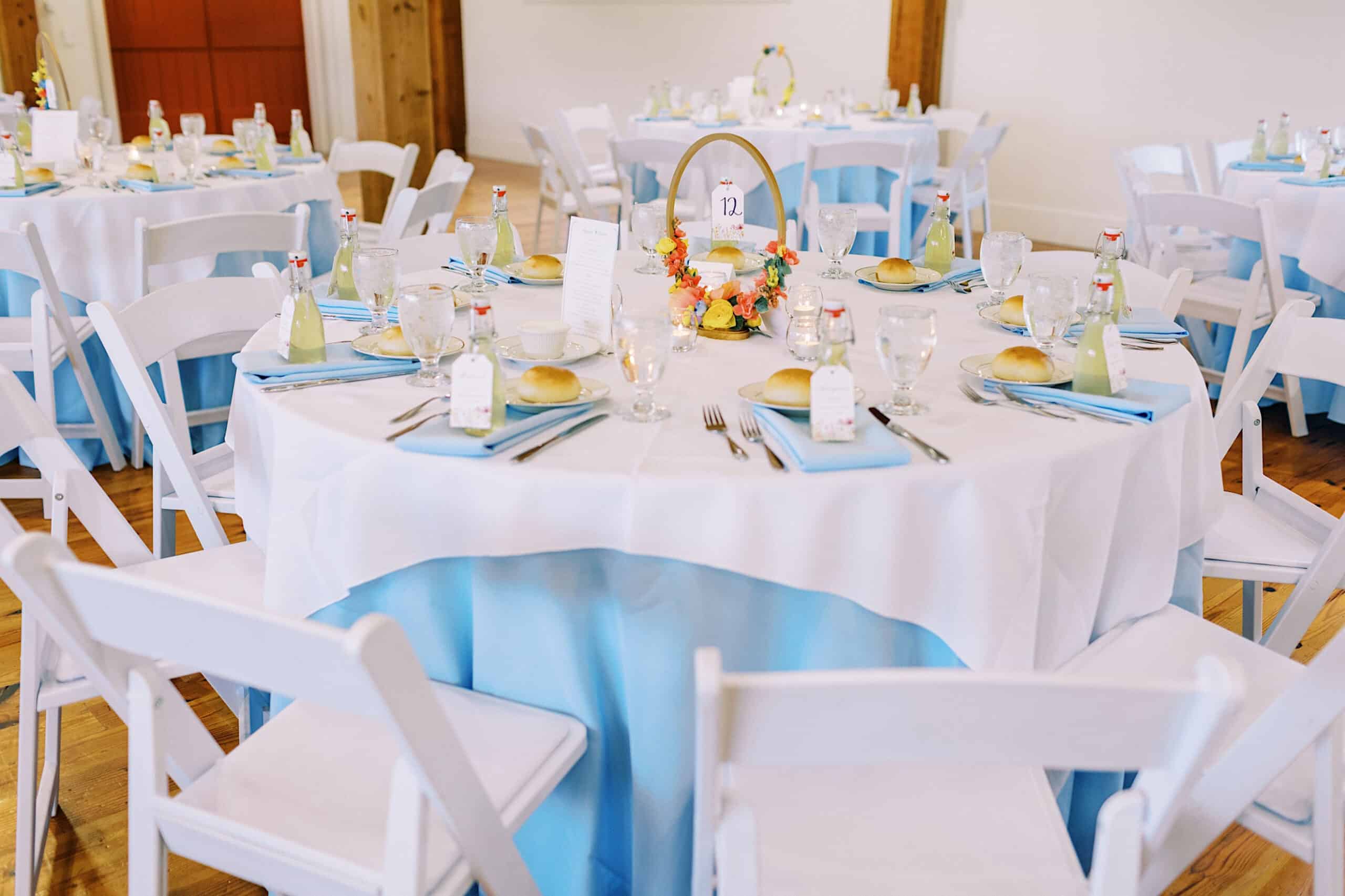 Round table set for a Summer Wedding at The High Point, with white tablecloth, light blue accents, place settings, bread rolls, glass bottles, and a floral centerpiece with a table number sign. White chairs surround the elegant setup.