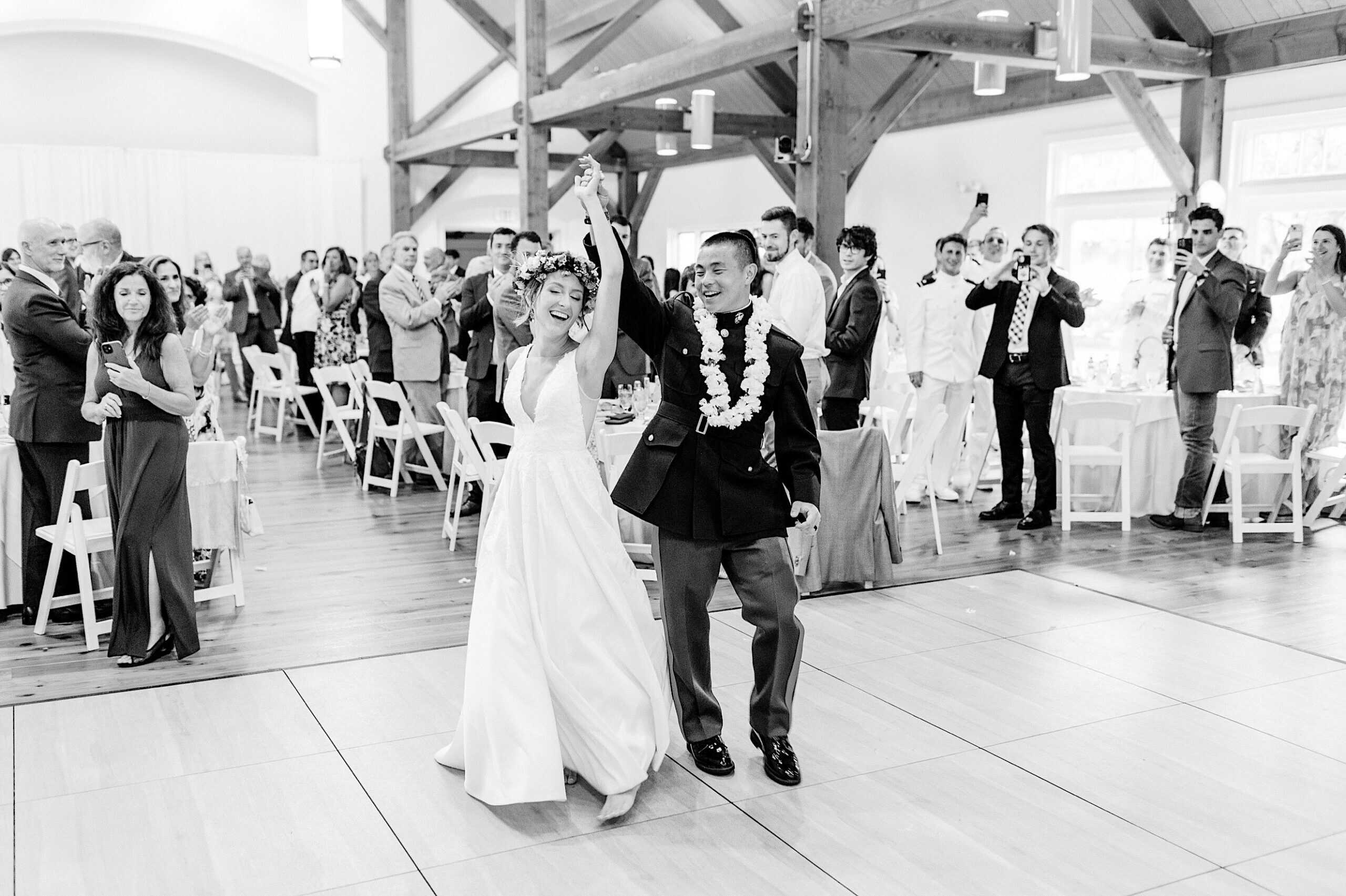 A bride and groom smile and raise their hands while entering their Summer Wedding at The High Point, surrounded by standing guests who are clapping and taking photos.